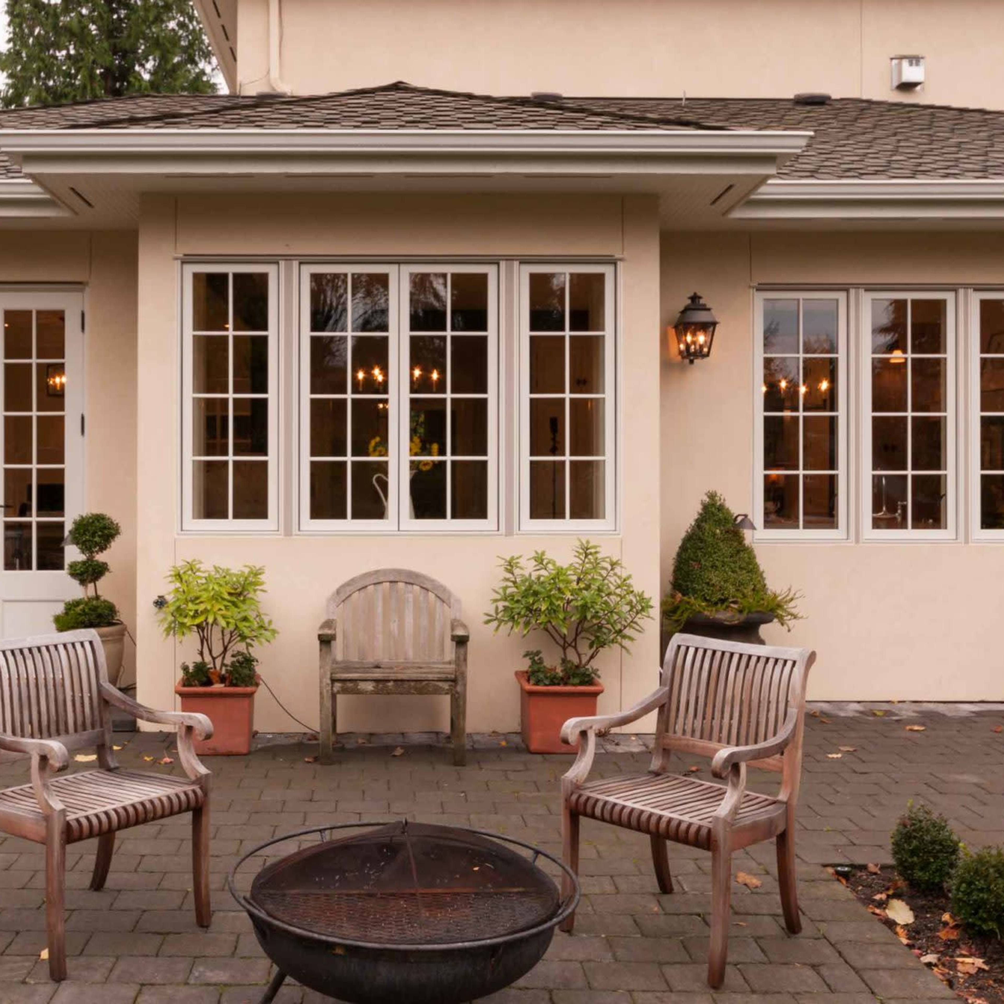 A residential patio area featuring two wooden chairs, a fire pit, and potted plants in front of a house with multiple windows and exterior lamps.