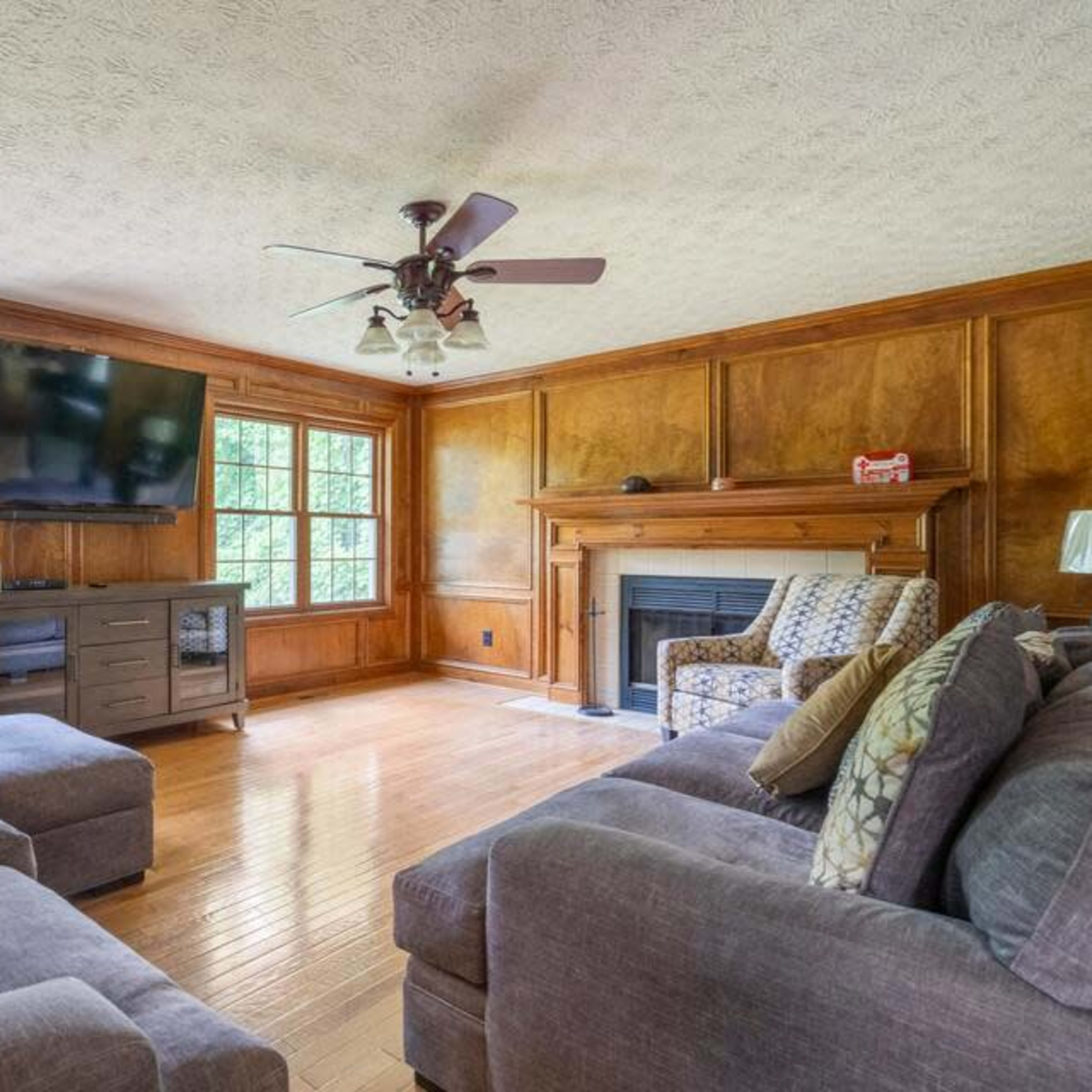 A living room features two sofas, a patterned armchair, a television mounted on the wall, and a wooden fireplace against a paneled backdrop.