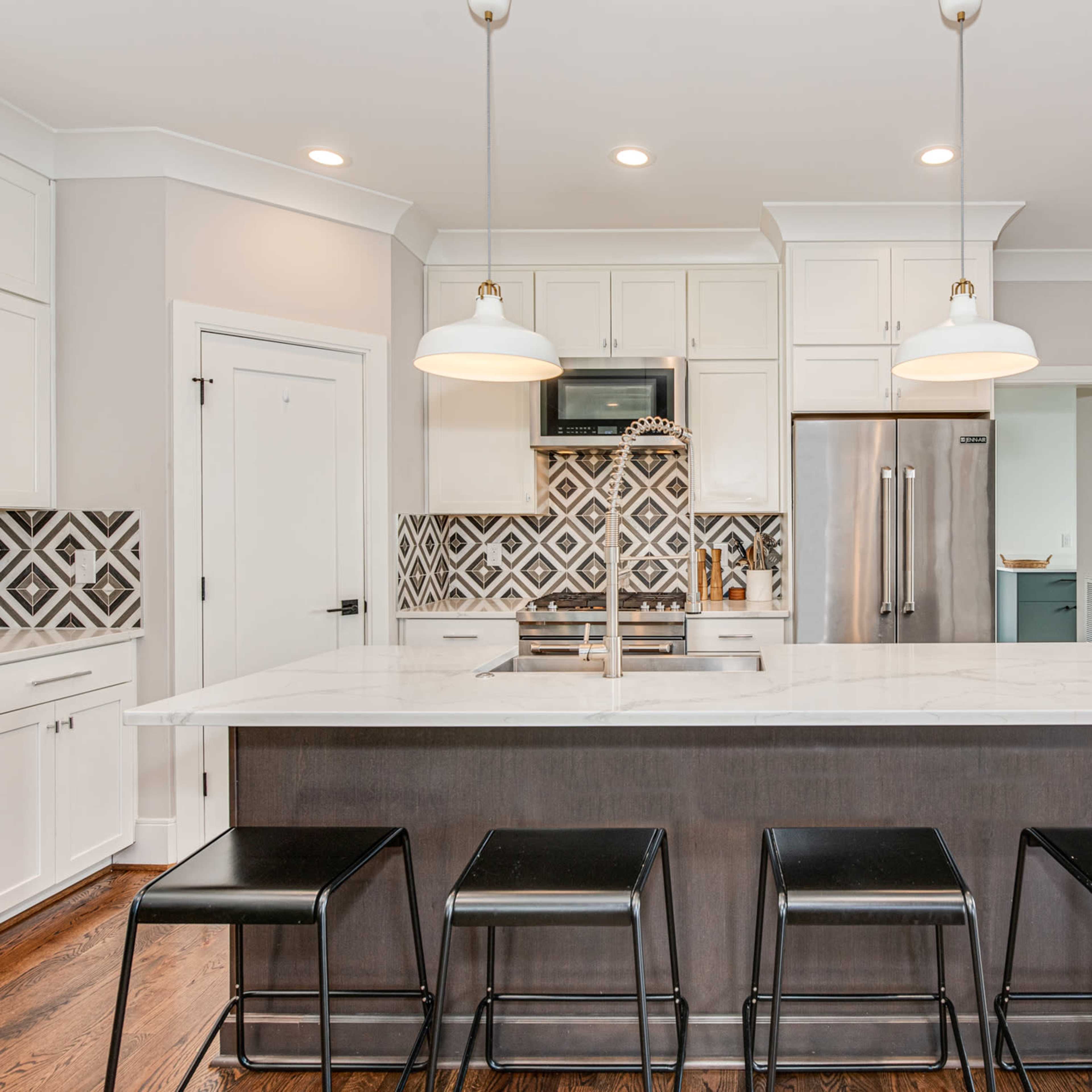A modern kitchen features a large island with black stools, white cabinetry, and a patterned backsplash.
