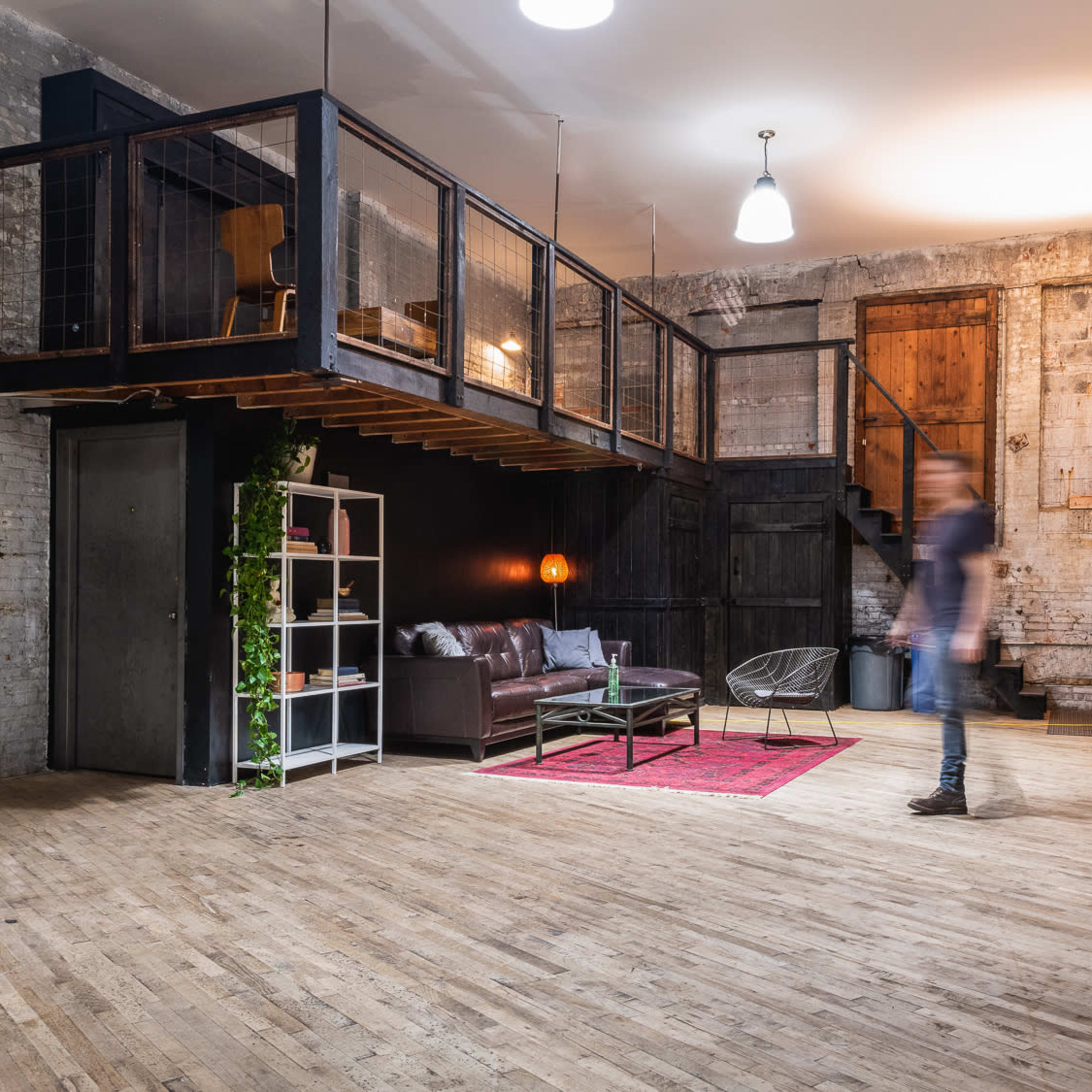 An industrial-style loft space with a wooden floor, a raised loft area, a modern seating area, and a mannequin near a bookshelf.