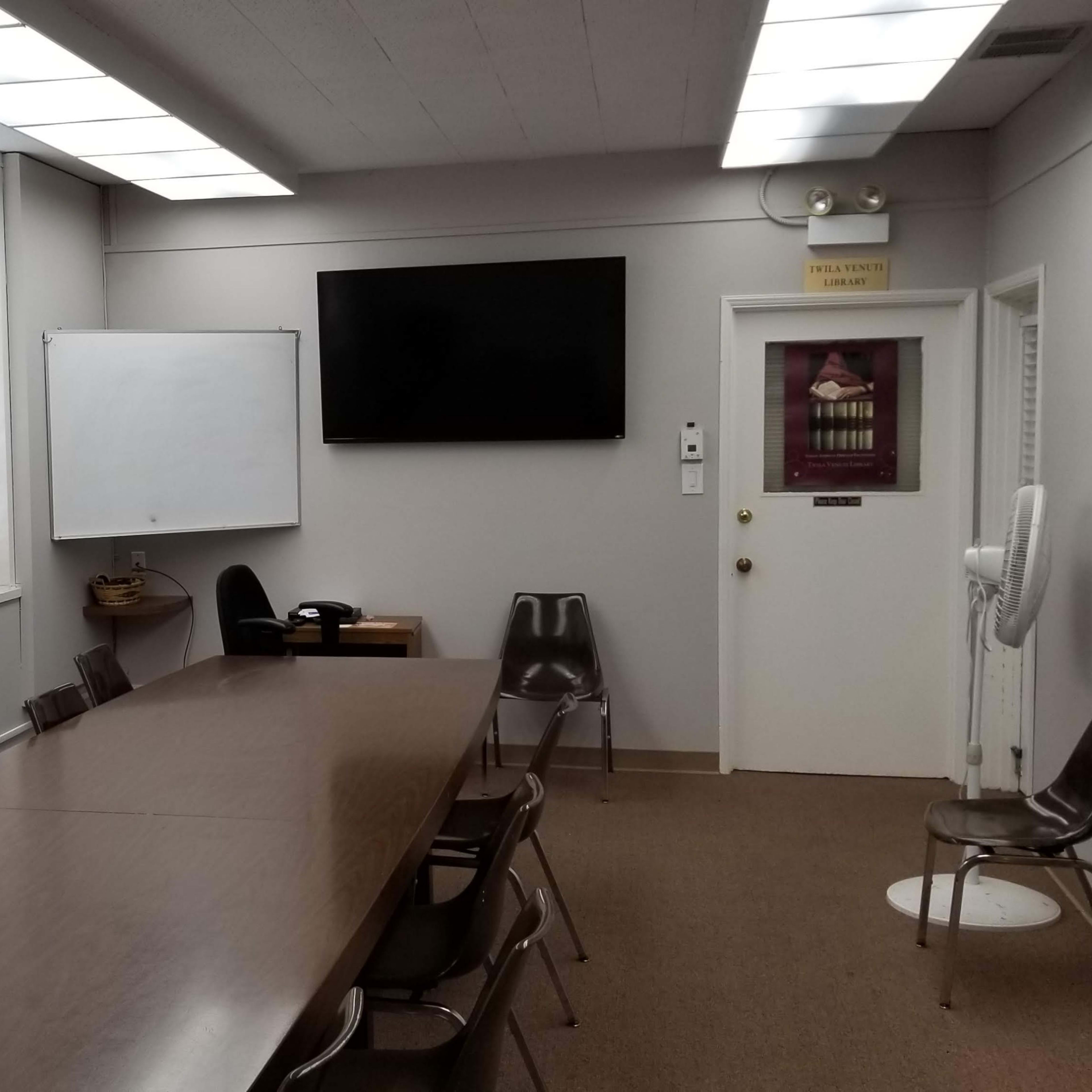 A conference room features a long wooden table, several chairs, a whiteboard, a television mounted on the wall, and framed paintings.