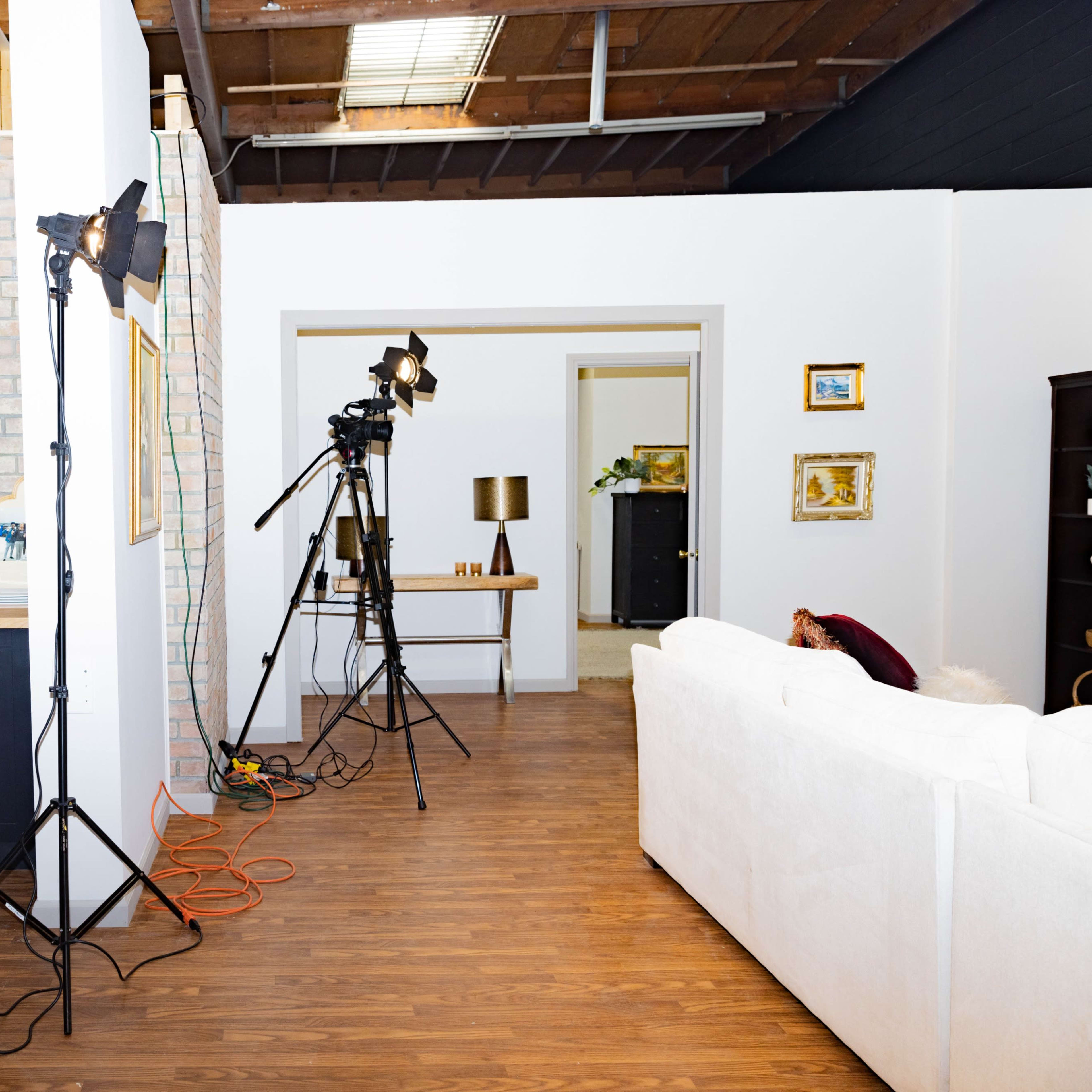 The scene features a studio set-up with a kitchenette, a white couch, and camera equipment positioned on tripods.
