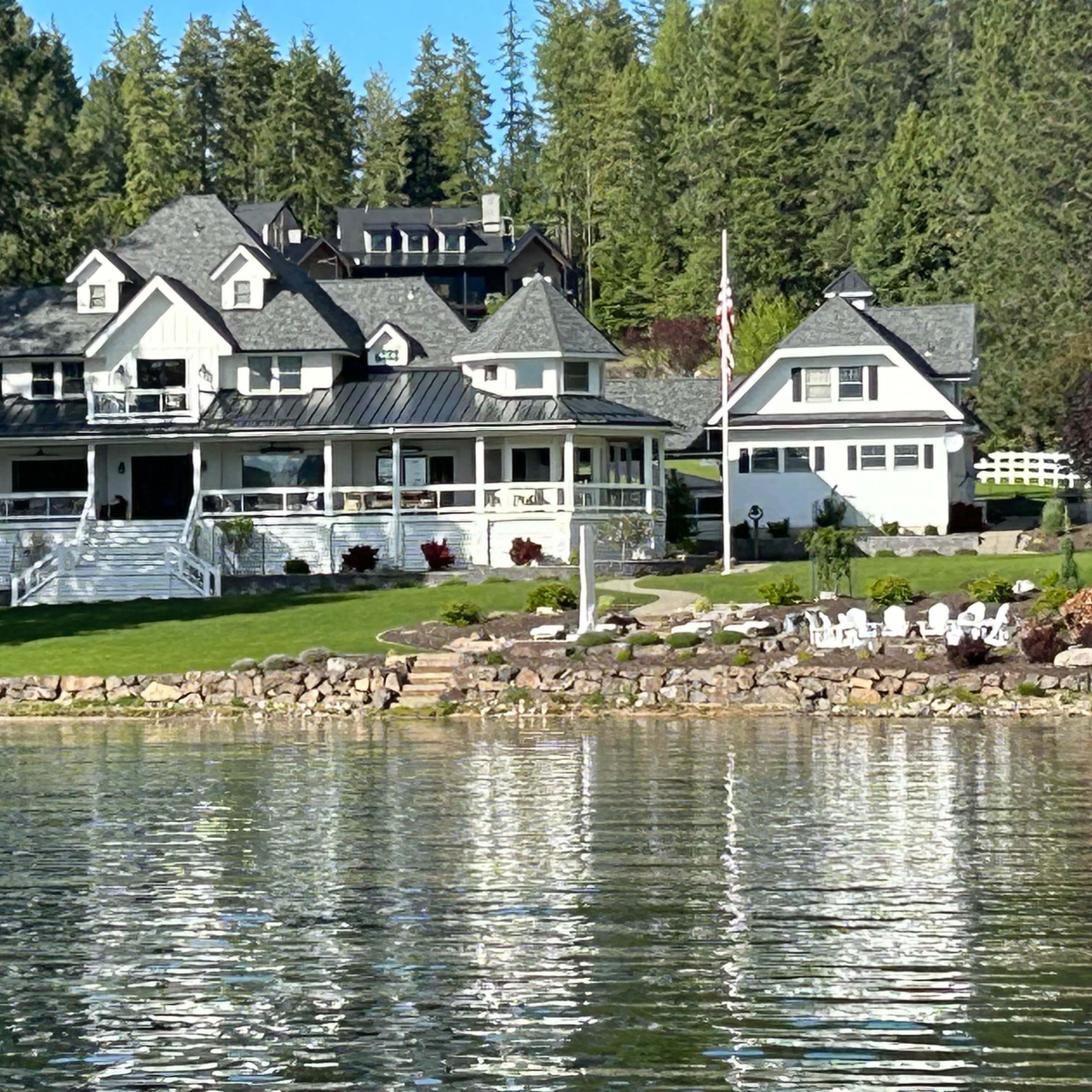 A large, multi-level white house with a wraparound porch and several smaller buildings on a shoreline, surrounded by trees and reflecting in calm water.