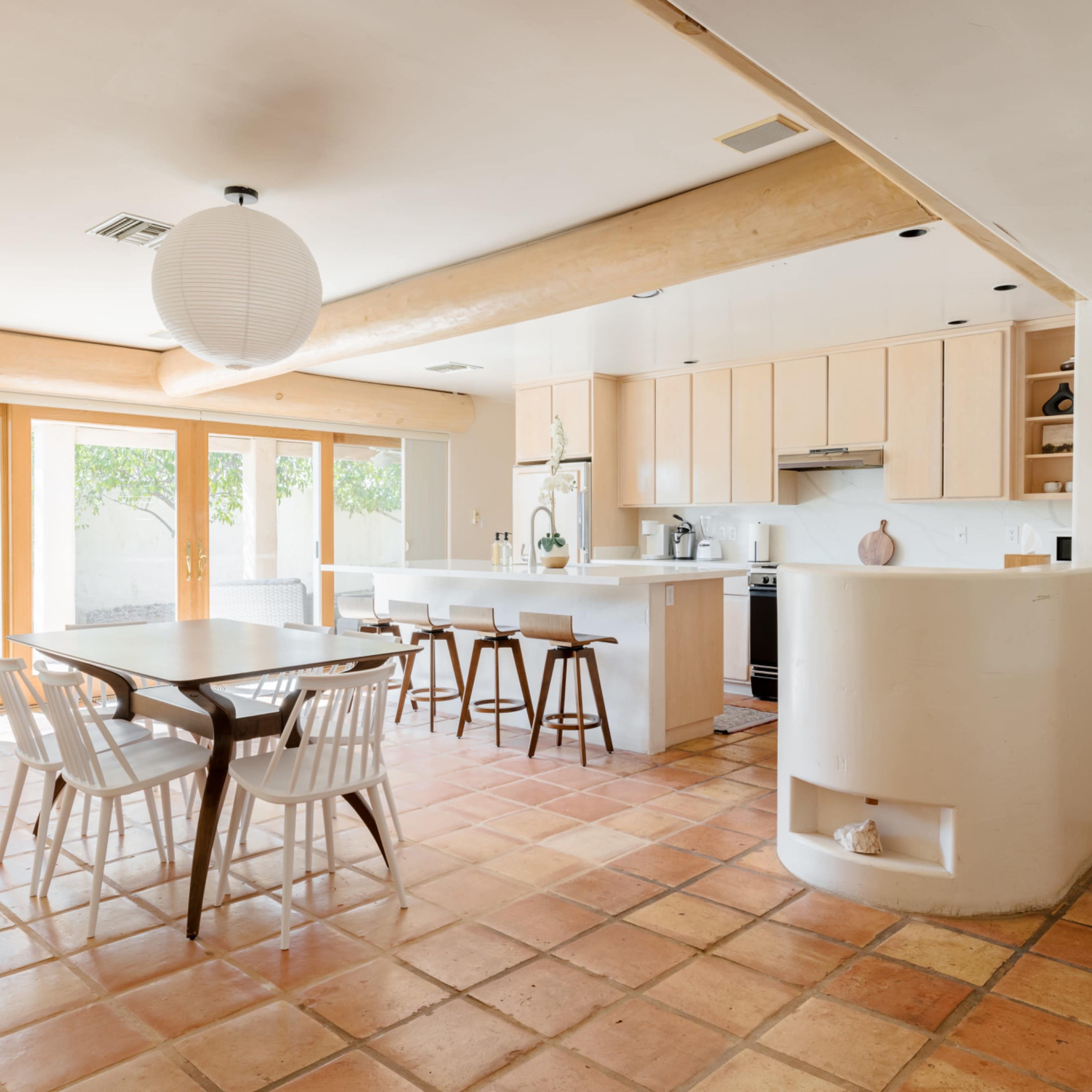A spacious kitchen and dining area features a table with white chairs, light wood cabinets, and large sliding glass doors leading to a garden.