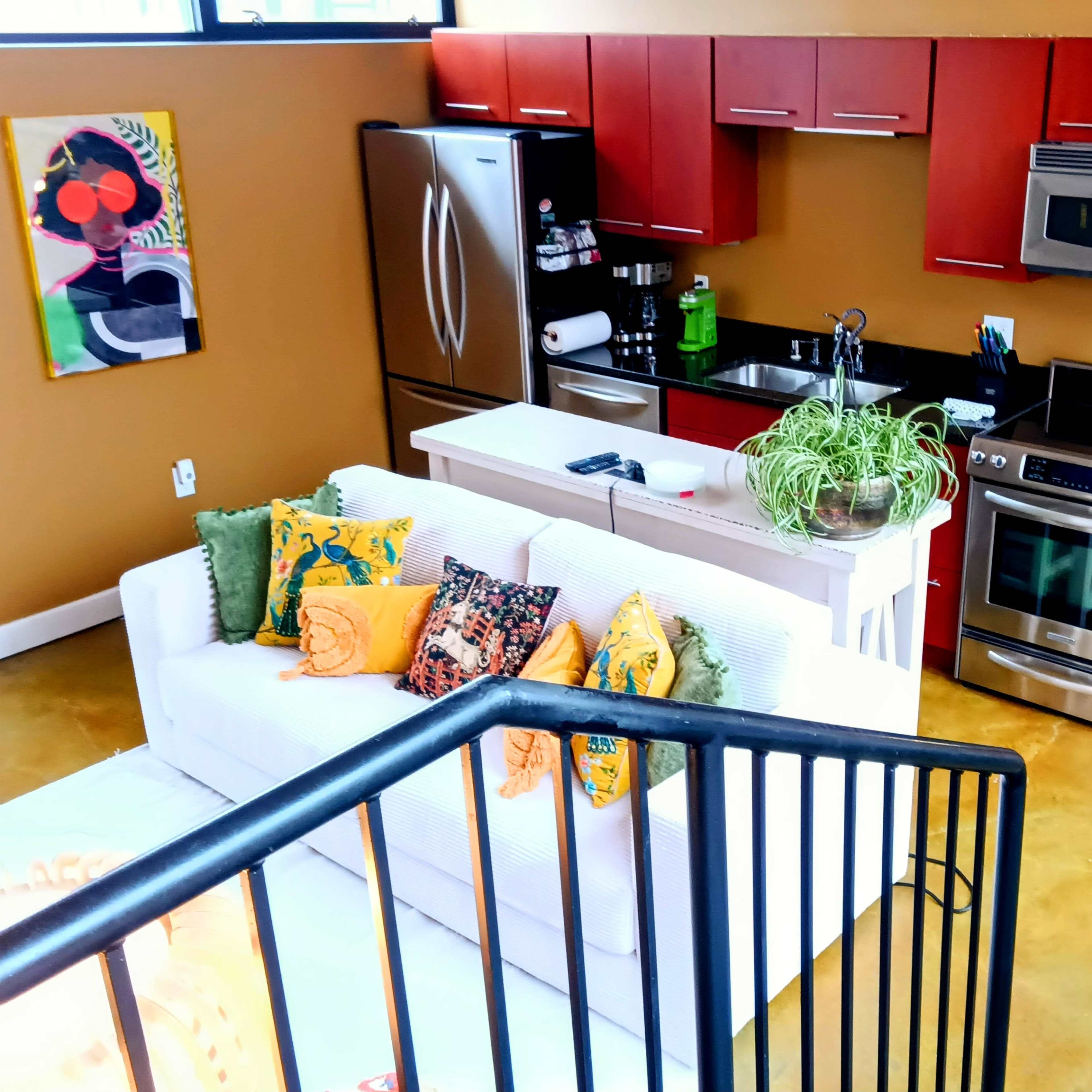 A modern kitchen with red cabinetry, stainless steel appliances, and a white couch decorated with colorful cushions.