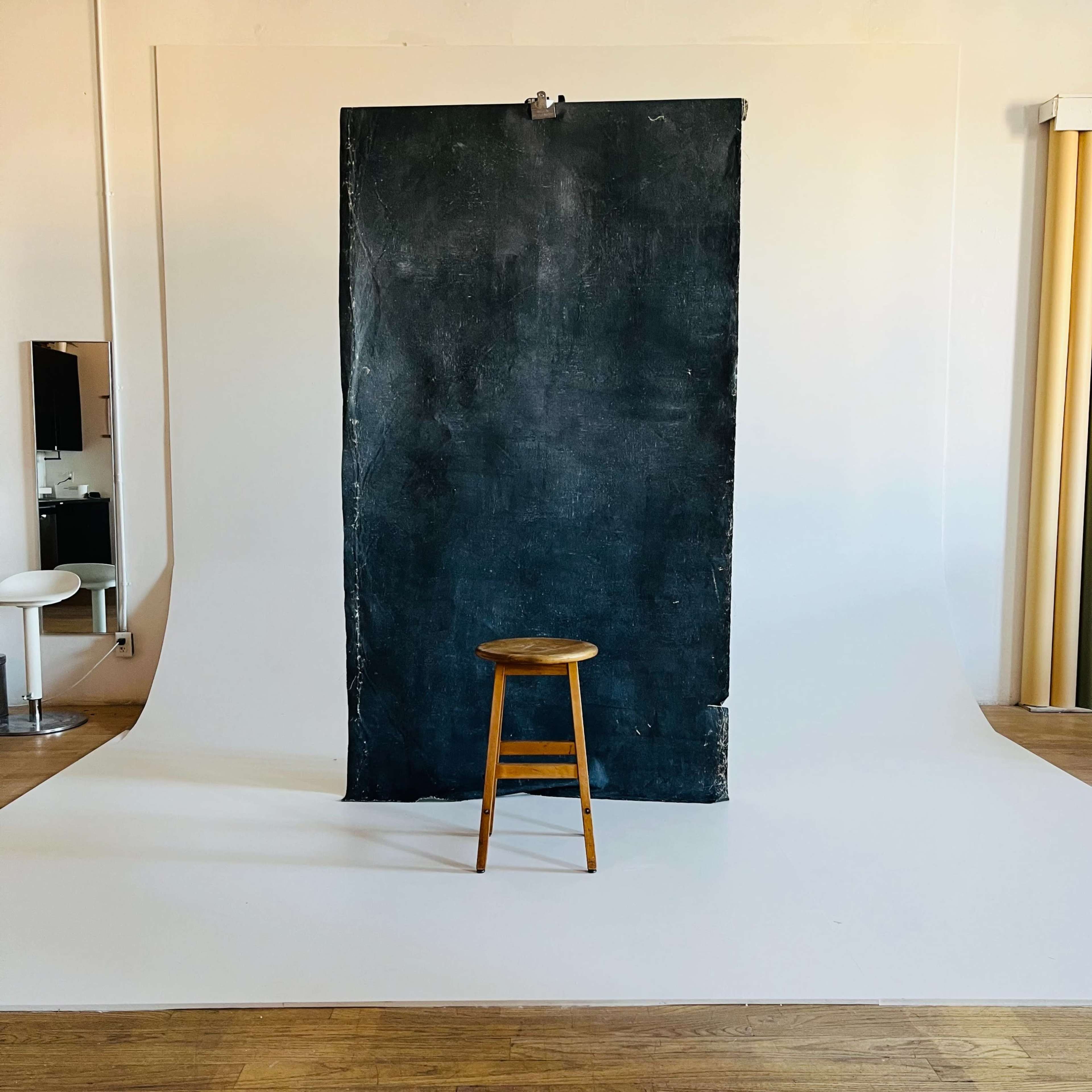 A wooden stool stands in front of a large, dark backdrop within a photo studio.