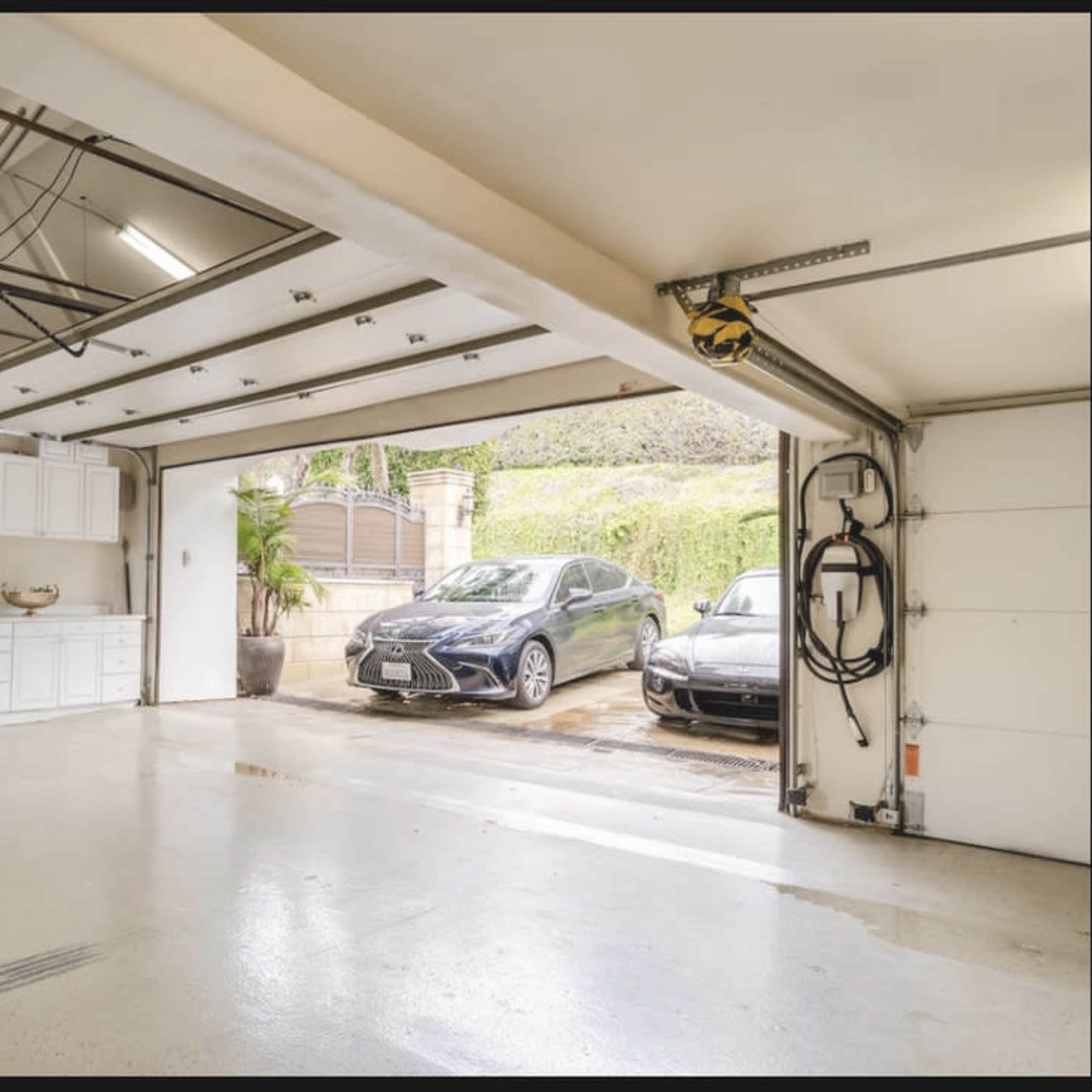 A spacious garage with polished concrete floors, featuring a wall of cabinets on one side and two cars parked in the driveway beyond the open doors.