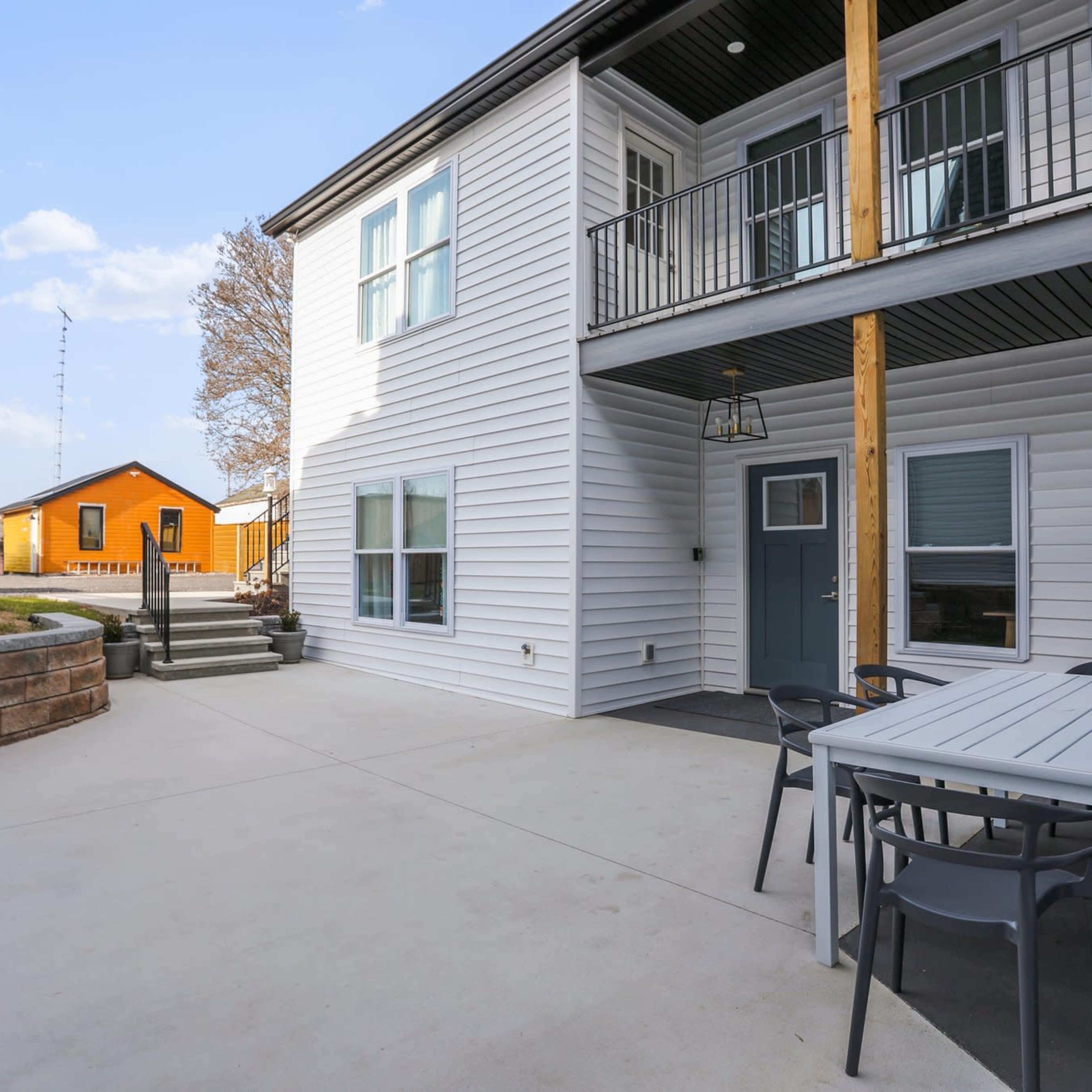 A modern two-story house with a spacious patio area, surrounded by a landscaped yard and a smaller colorful building in the background.