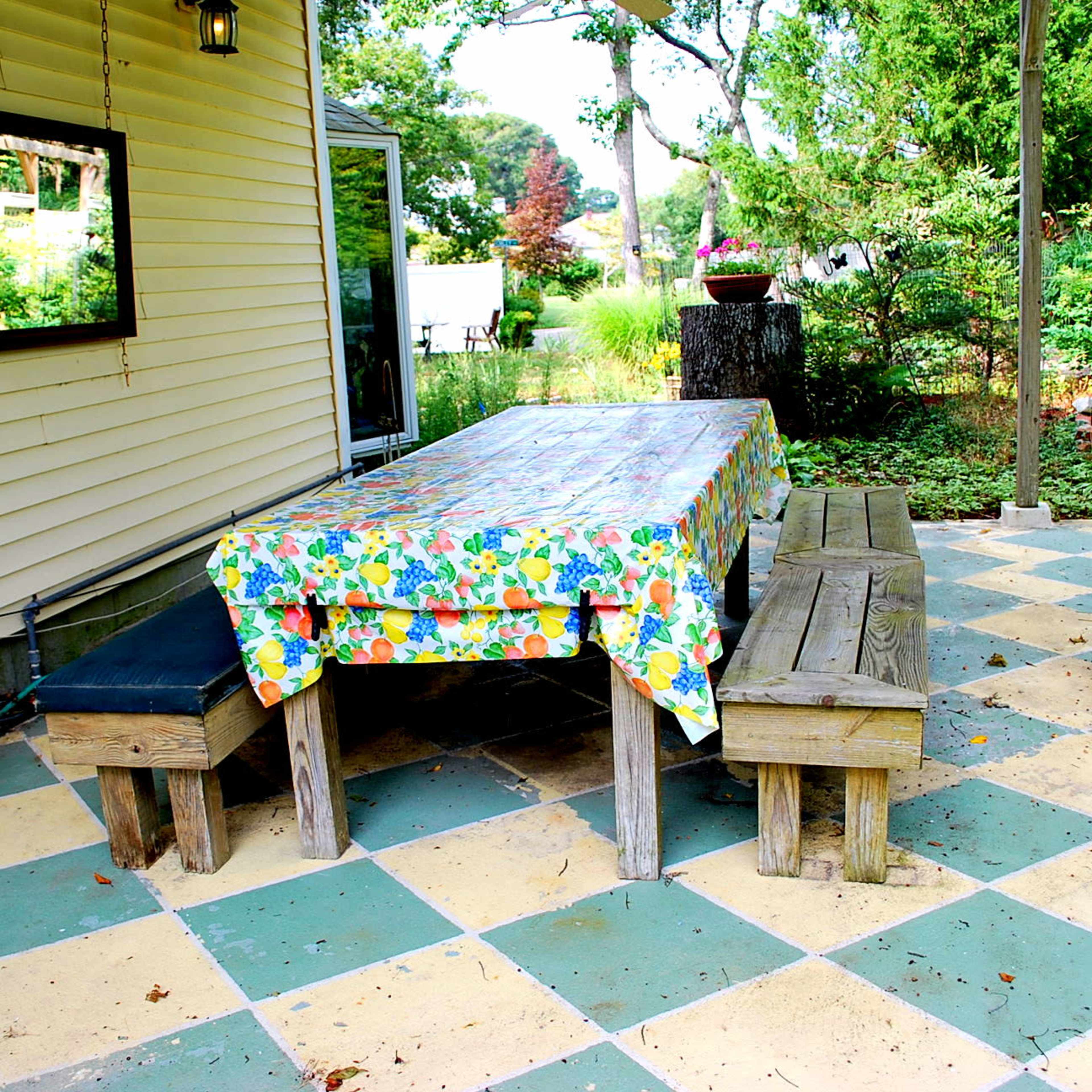 A wooden table covered with a colorful tablecloth is set in a garden area alongside two wooden benches and a patio featuring a checkerboard pattern.