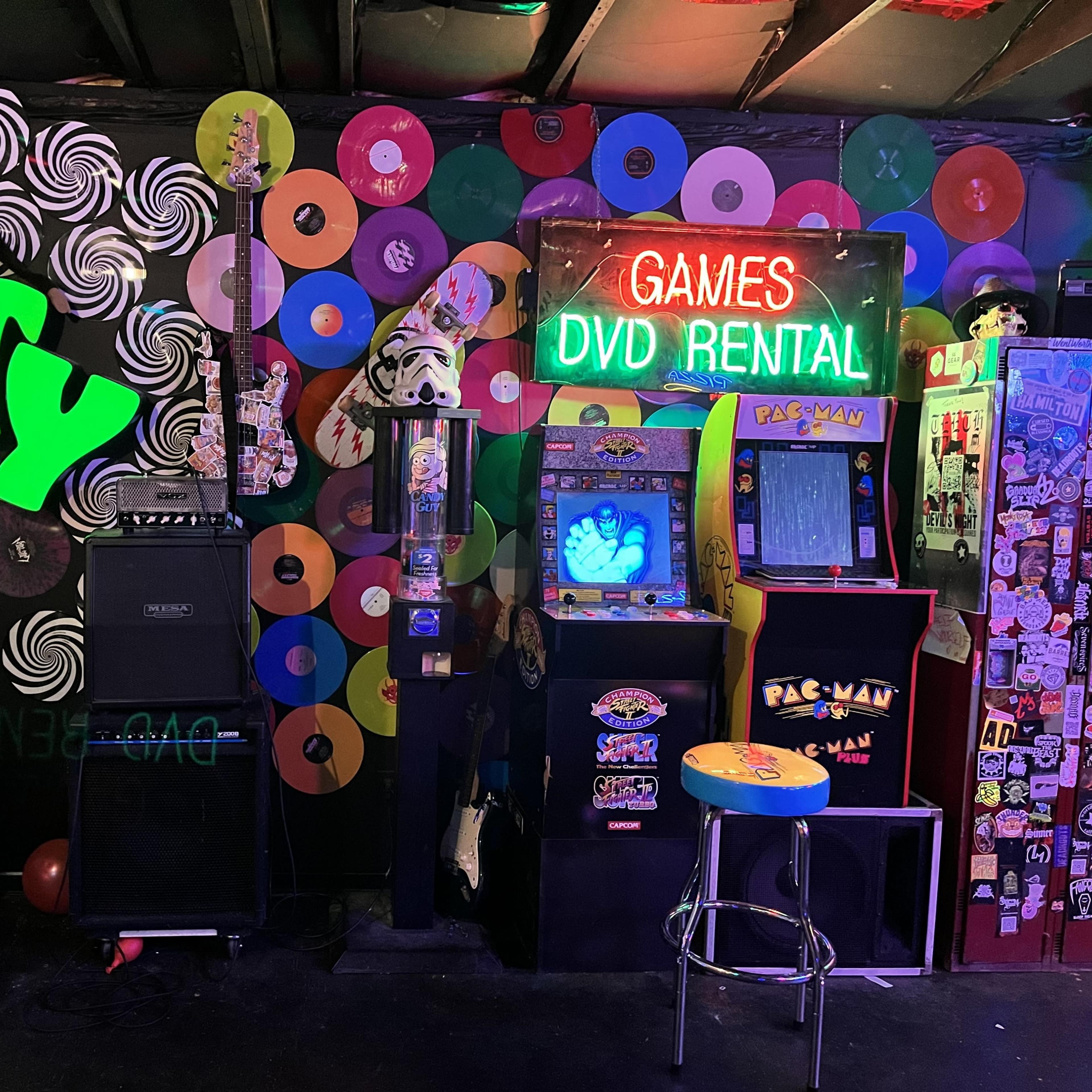 A vibrant arcade setup with a colorful wall of vinyl records, an arcade machine, and a small stage area with musical instruments.