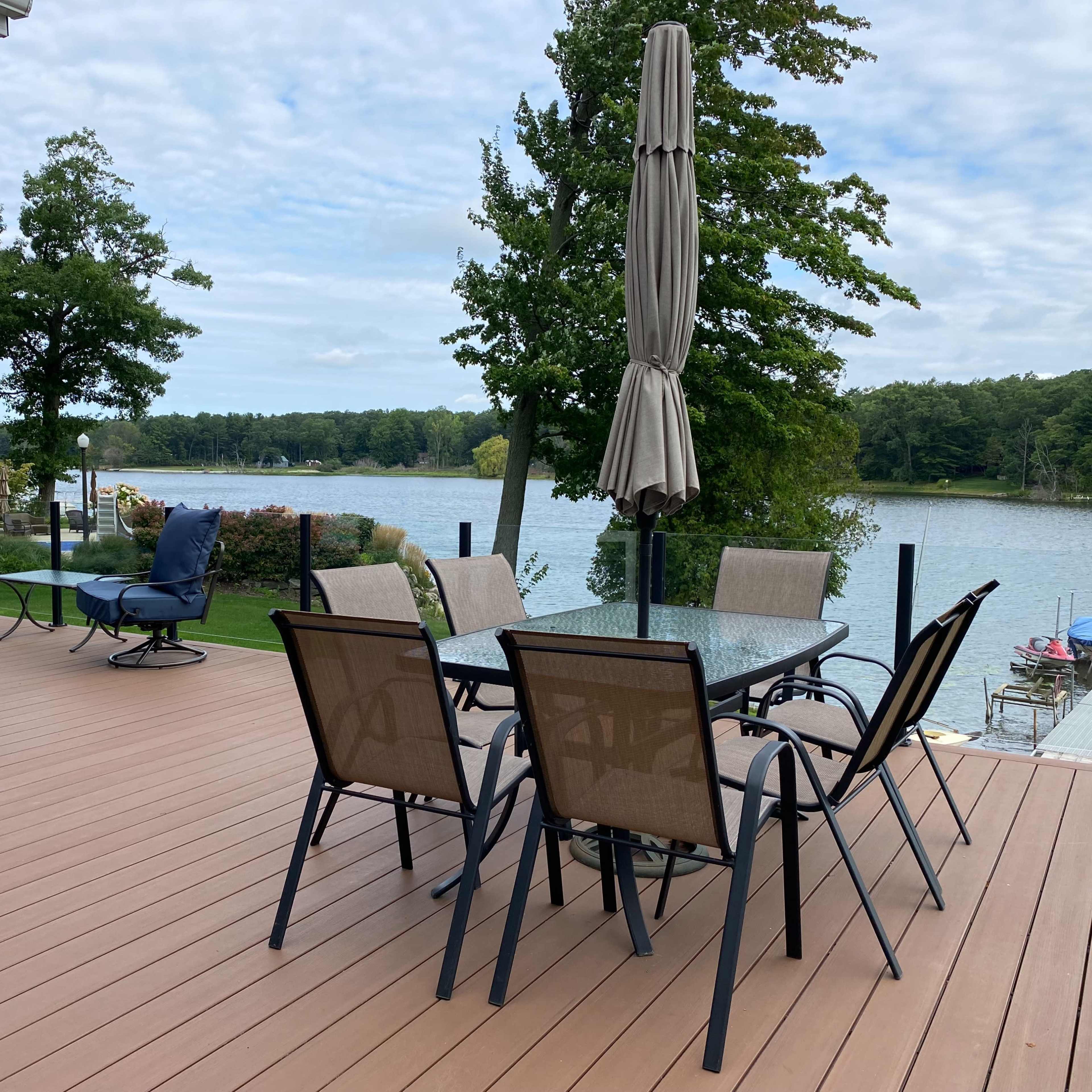 A wooden deck features a dining table with chairs, surrounded by lounge seating and overlooking a lake with boats and greenery in the background.