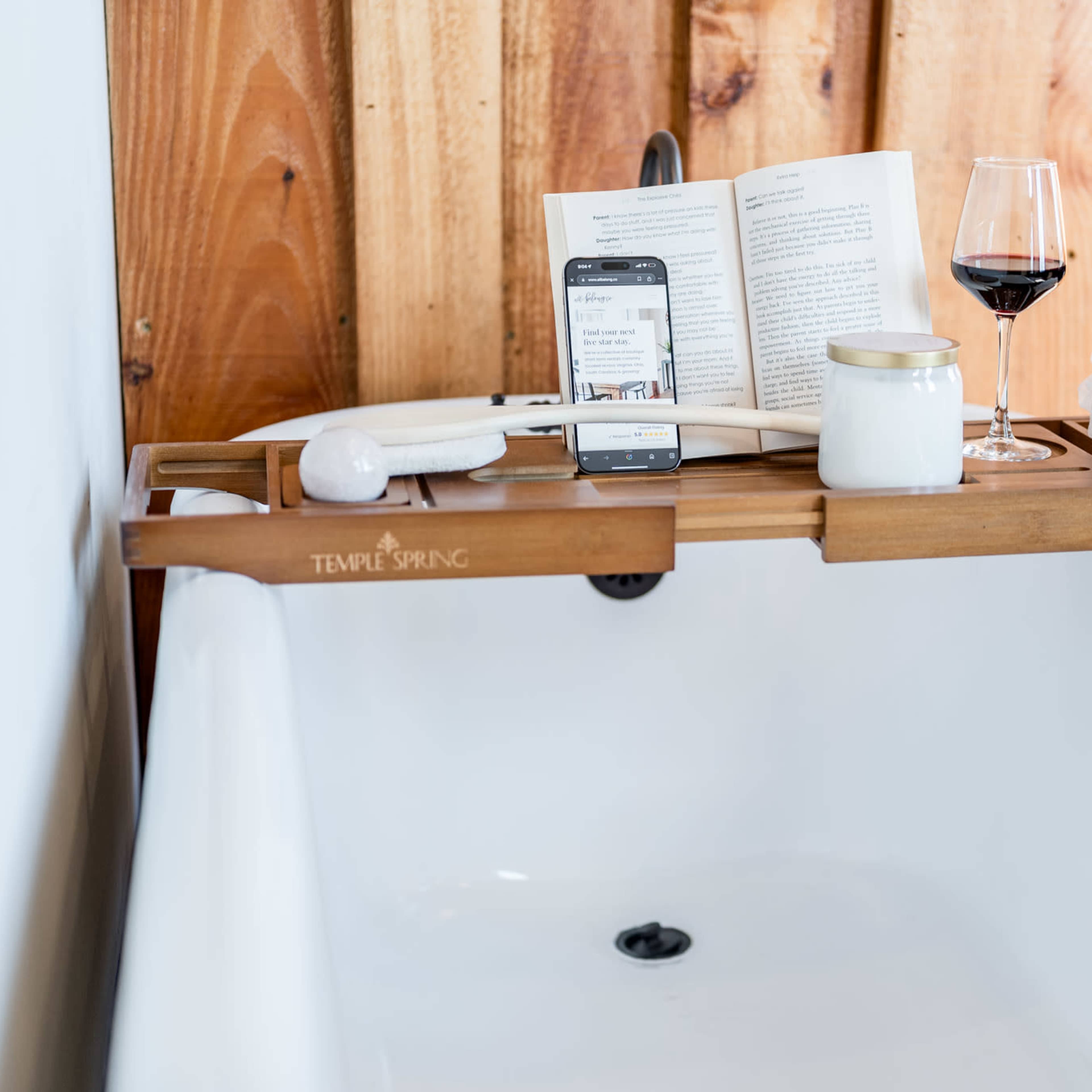 A wooden bathtub caddy holds a book, a glass of red wine, a jar, a loofah, and a smartphone over the edge of a white tub, against a backdrop of wooden walls.