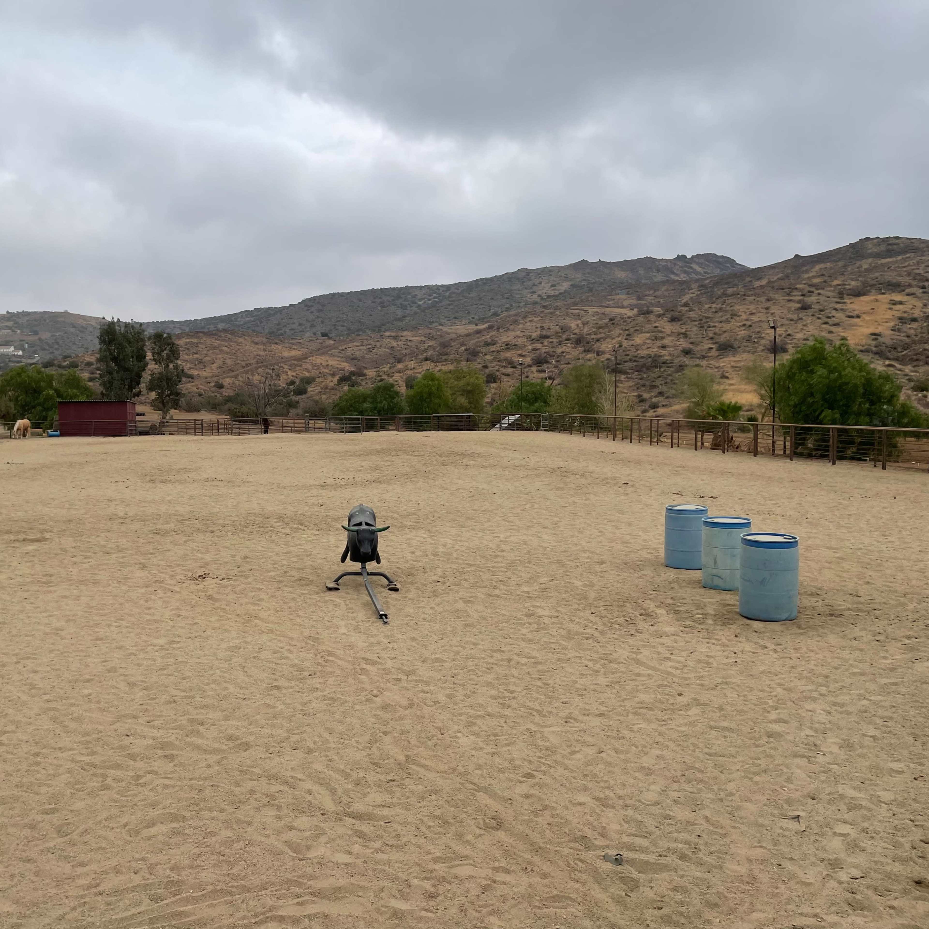 A sandy outdoor arena is surrounded by hills and sparse vegetation, featuring a riding apparatus and a few blue barrels.