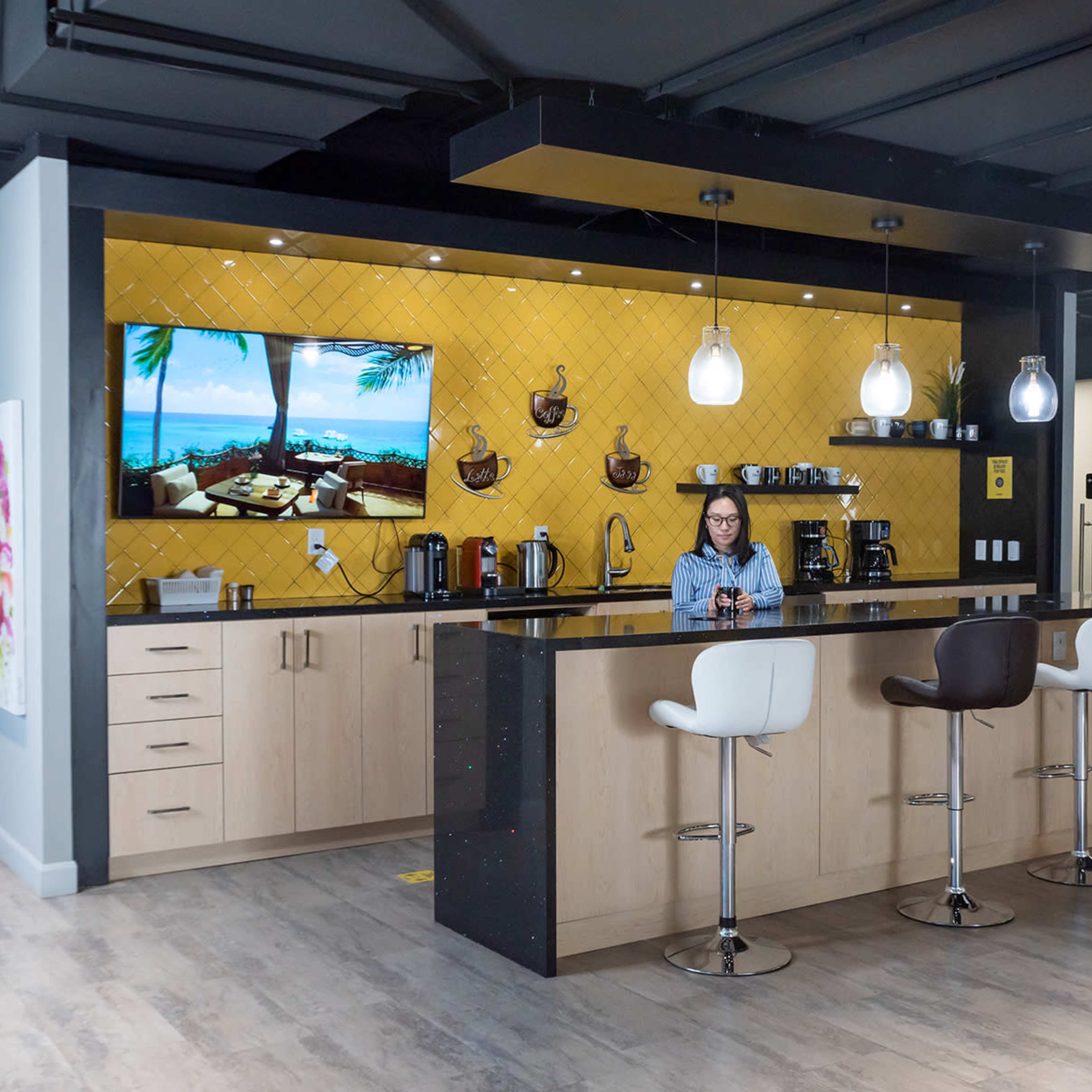 A modern kitchen area features a woman standing at a black countertop with bar stools, alongside a wall-mounted television displaying a tropical scene.