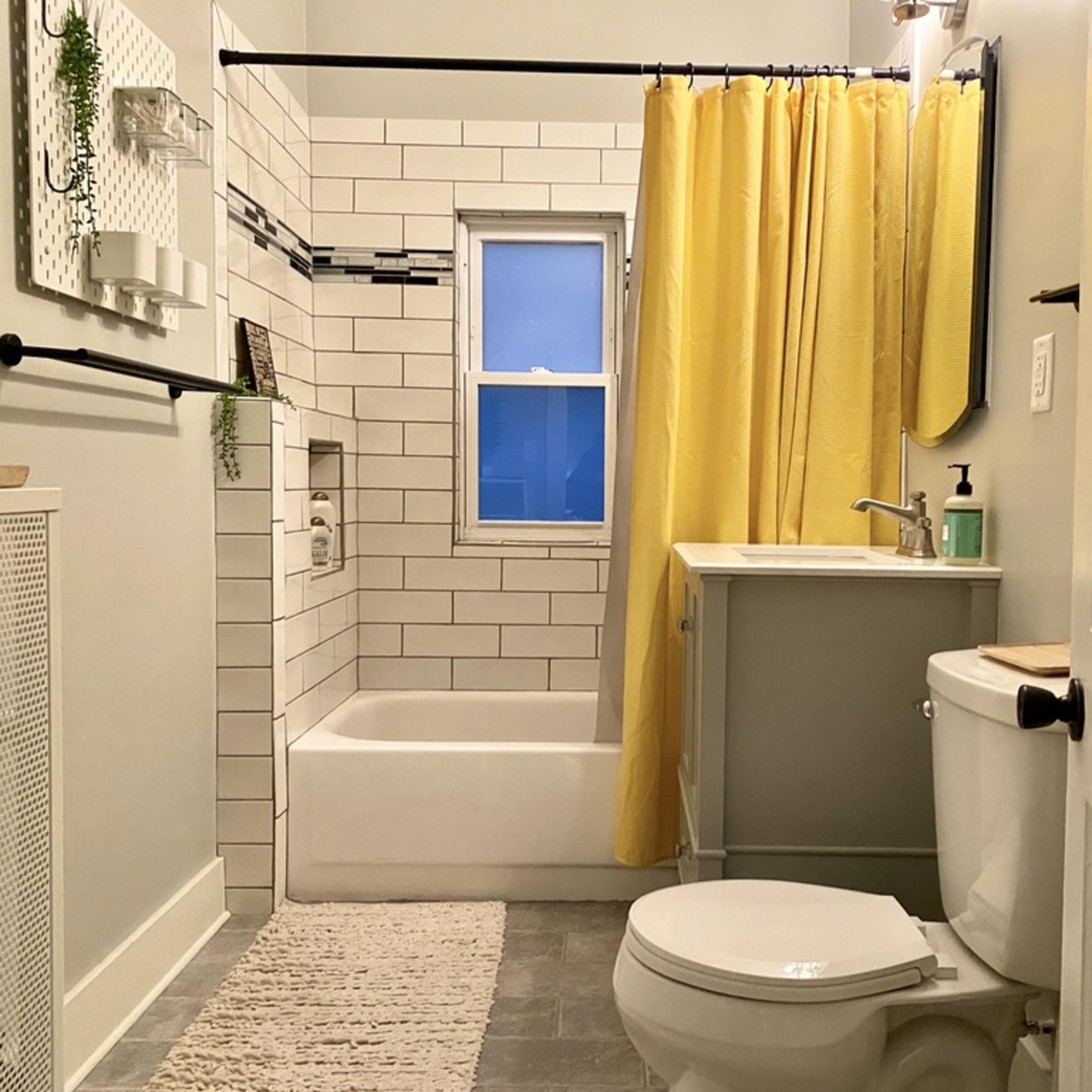 A bathroom featuring a tub with a shower curtain, a wall-mounted mirror, a sink with a cabinet, and neutral-colored tiles on the floor and walls.