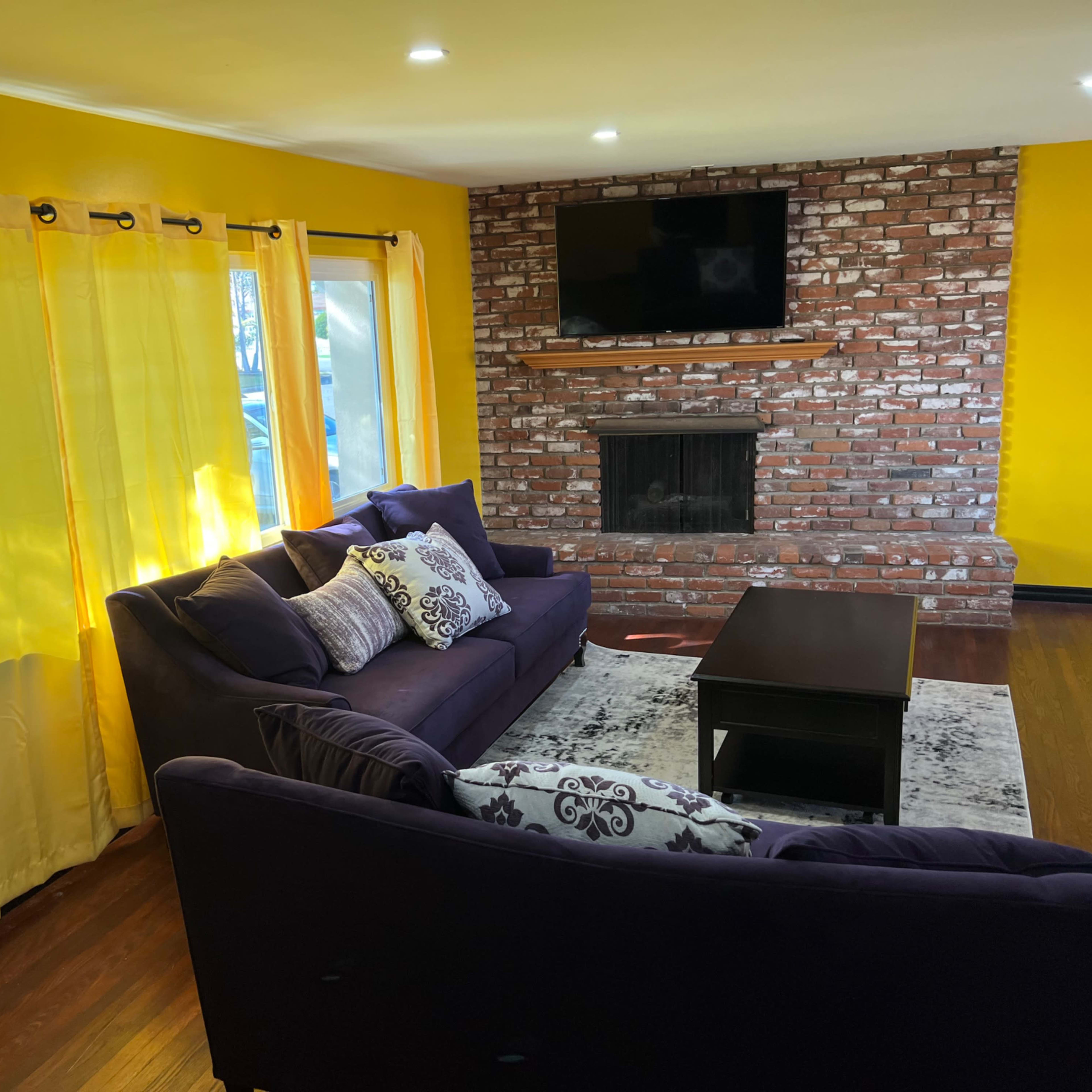 The living room features yellow walls, a brick fireplace, a black coffee table, and purple sofas with decorative pillows.