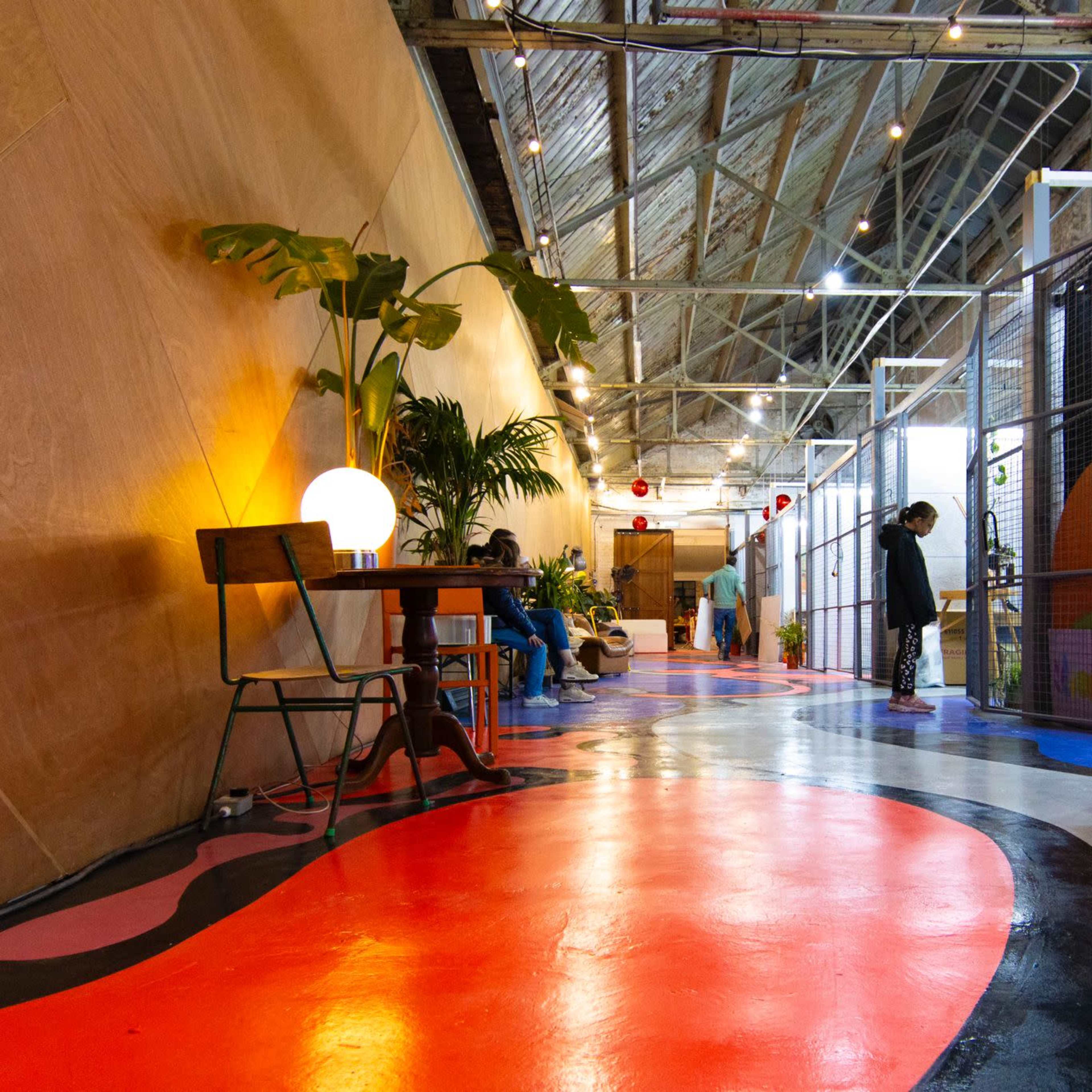 A colorful hallway with a mix of painted floors and plants, featuring tables and signs in a modern indoor space.