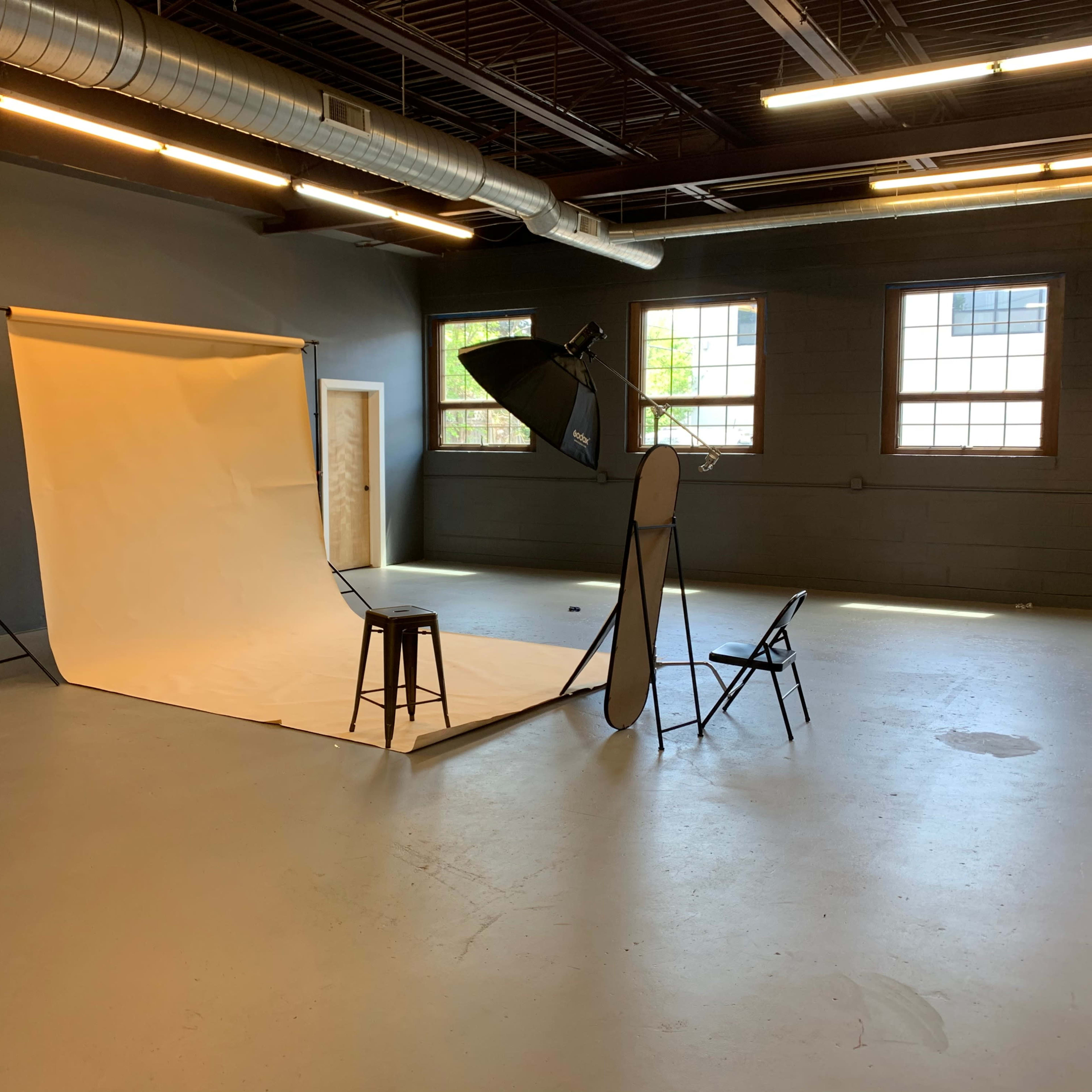 A photography studio setup with a backdrop, lighting equipment, a stool, and chairs arranged on a concrete floor.