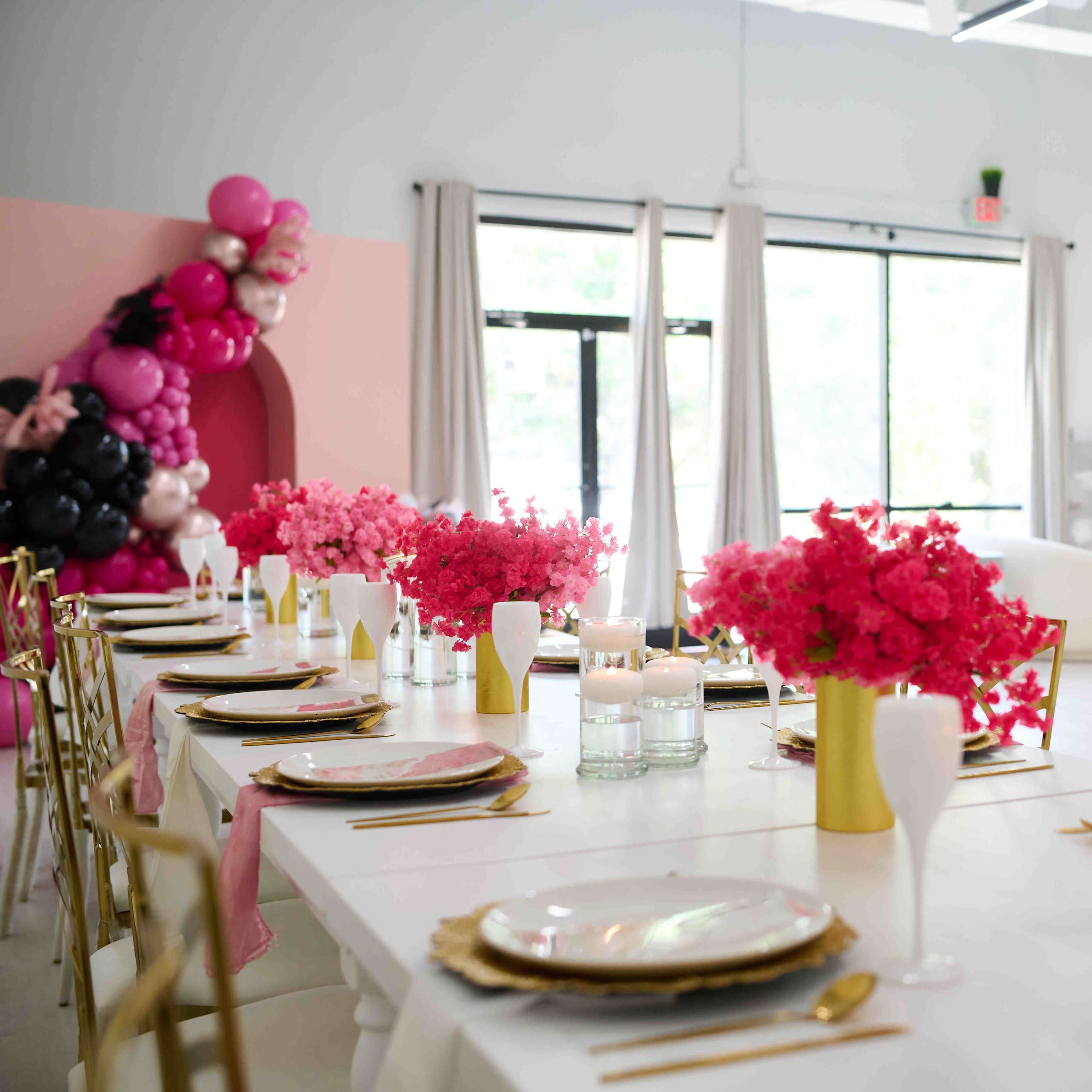 A long dining table adorned with pink flowers and candles sits in a brightly lit room decorated with balloons and modern furnishings.