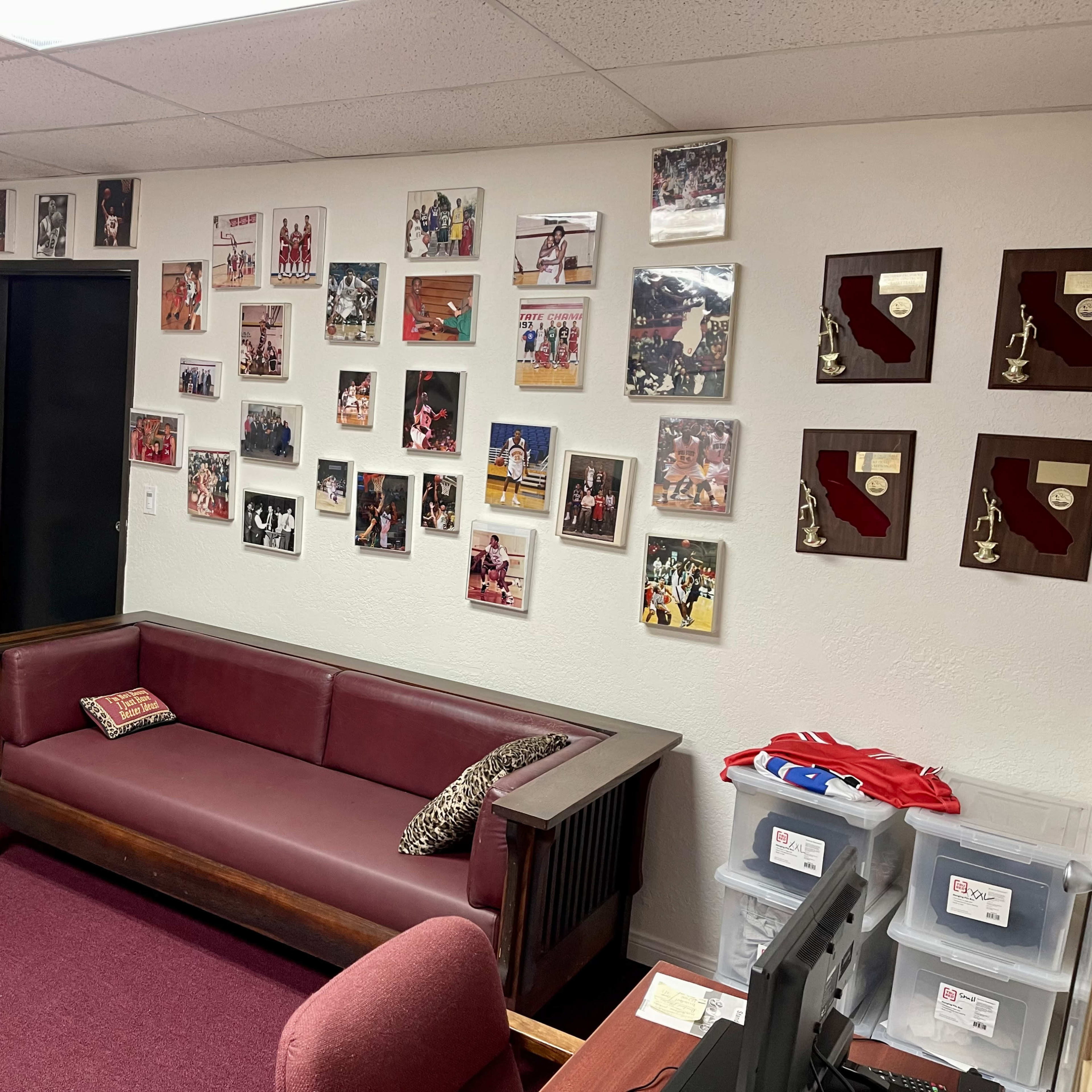 An office space with a maroon carpet, two wooden couches, a desk with a chair, and walls decorated with framed photos and awards related to sports.