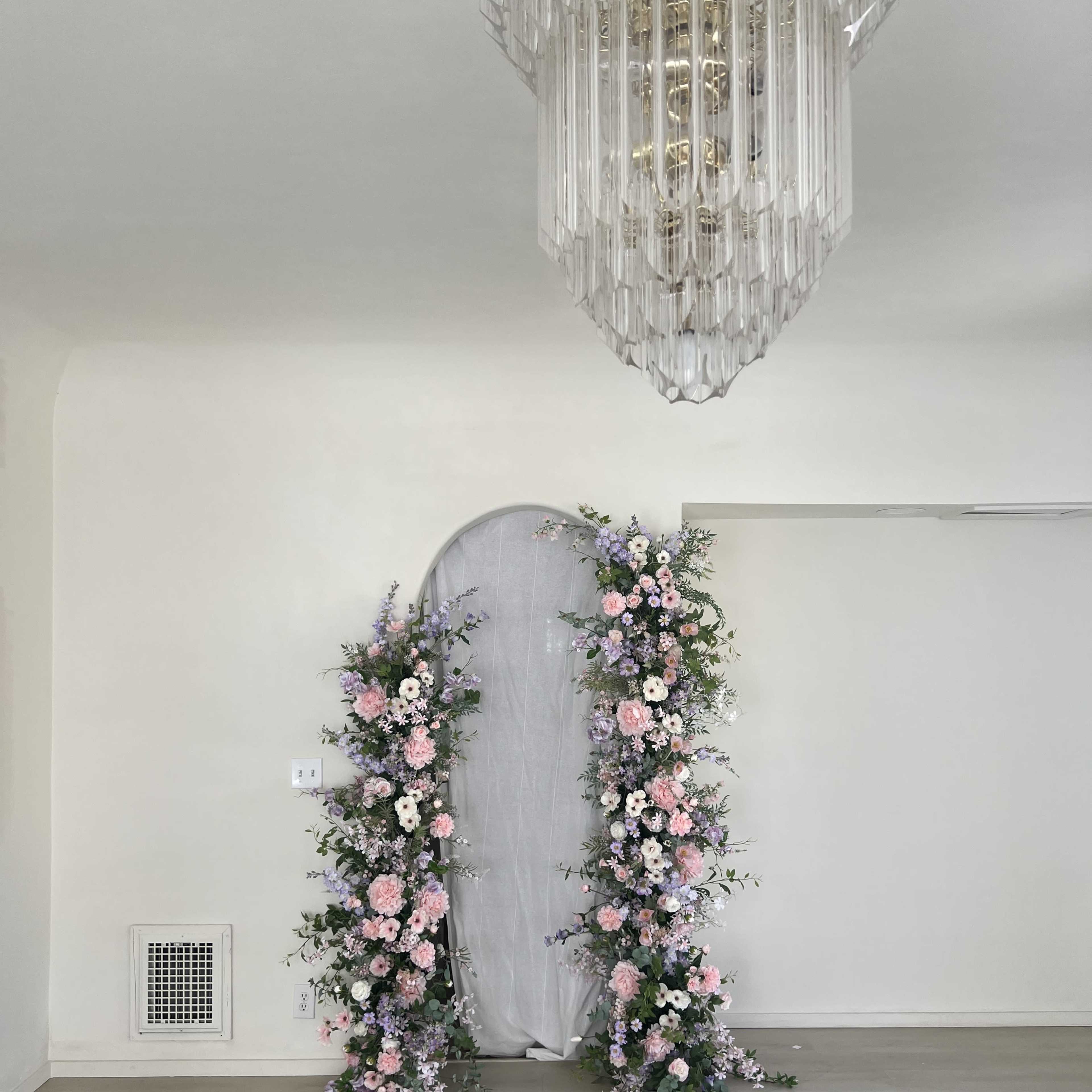 A floral arch adorned with pink and purple flowers in front of a light gray curtain, beneath a geometric crystal chandelier in a bright room.