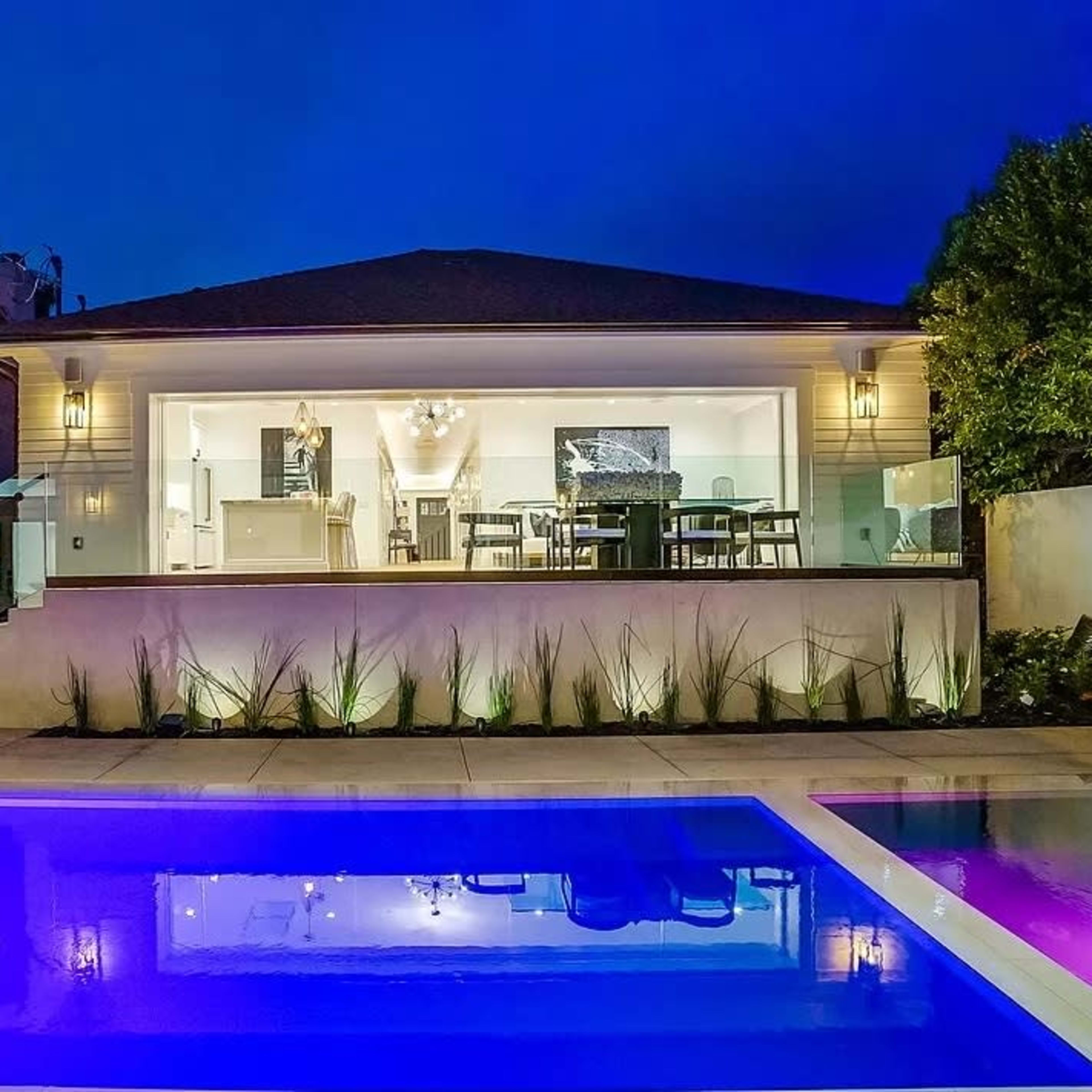 A modern house with large glass doors overlooks a swimming pool, illuminated by blue lights, in a night setting.