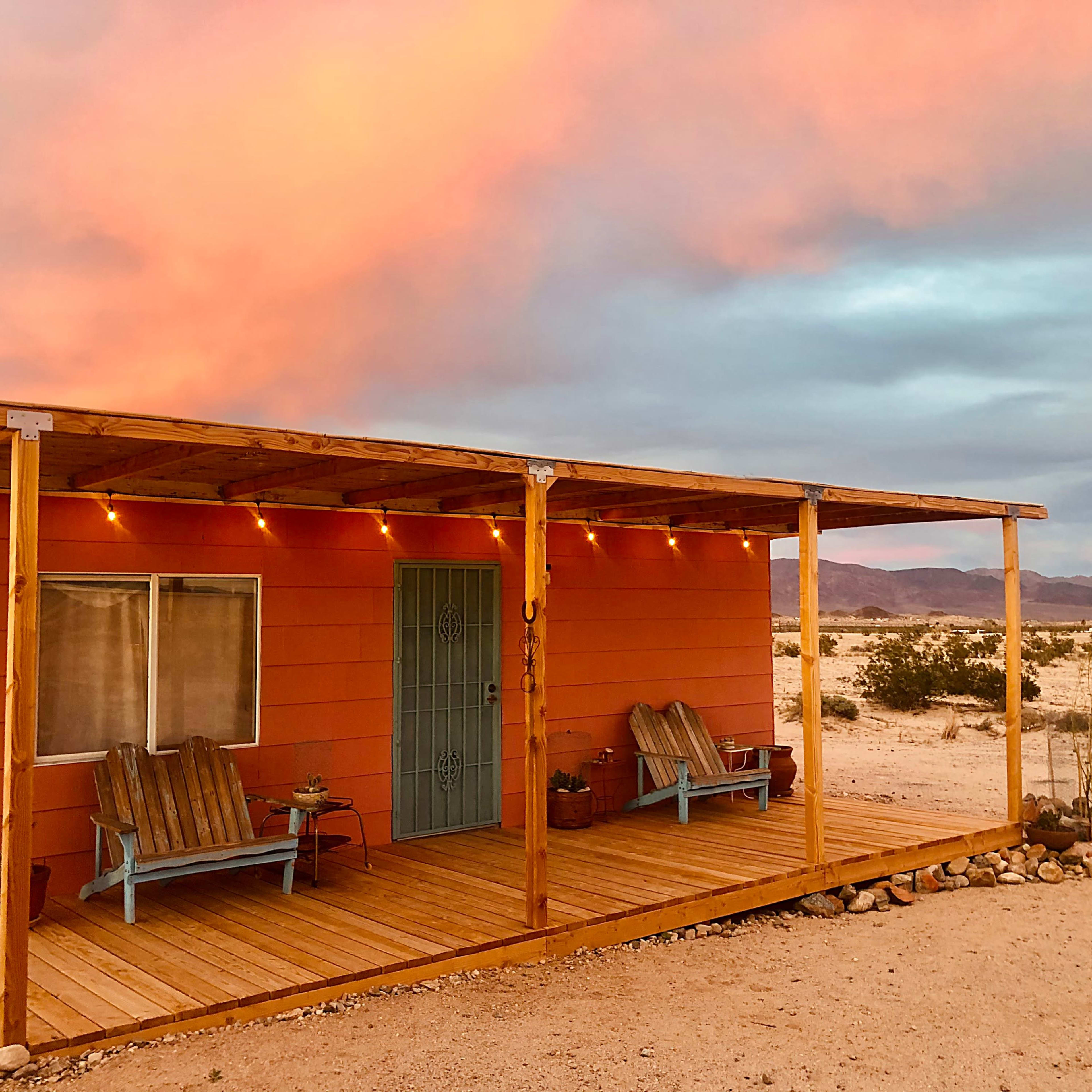 A small orange cabin with a wooden porch and blue chairs sits under a colorful sky in a desert landscape.
