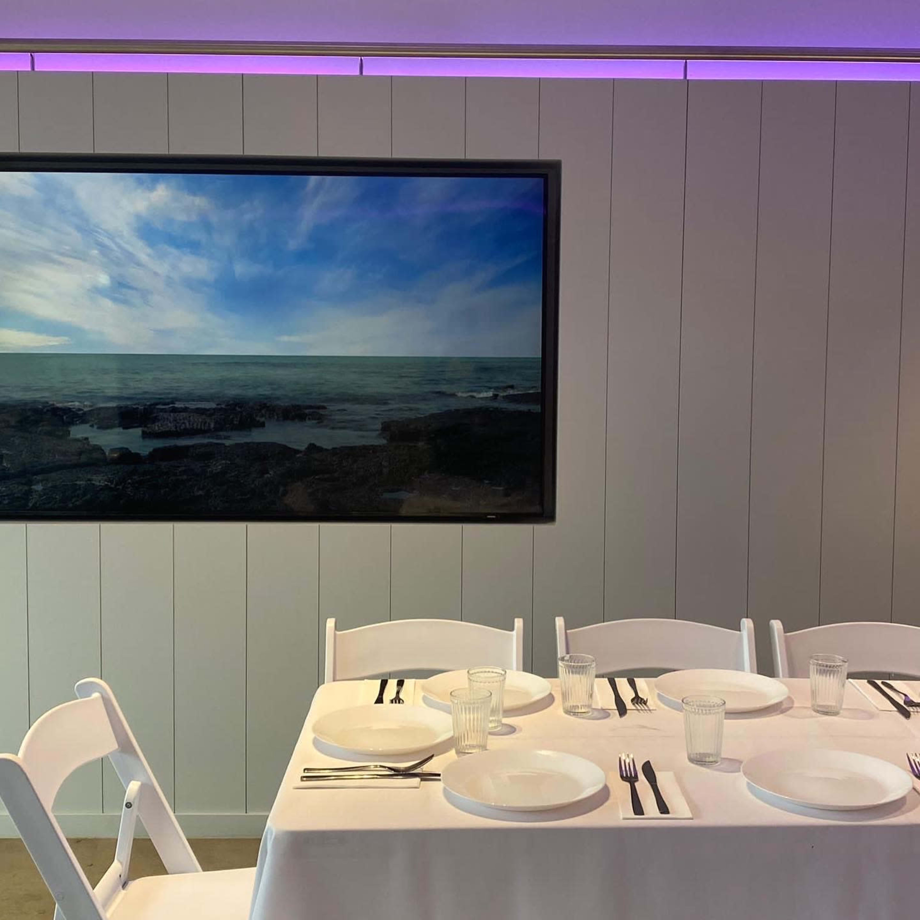 A dining table is set with white plates and utensils in front of a large framed landscape painting, with purple lighting above.