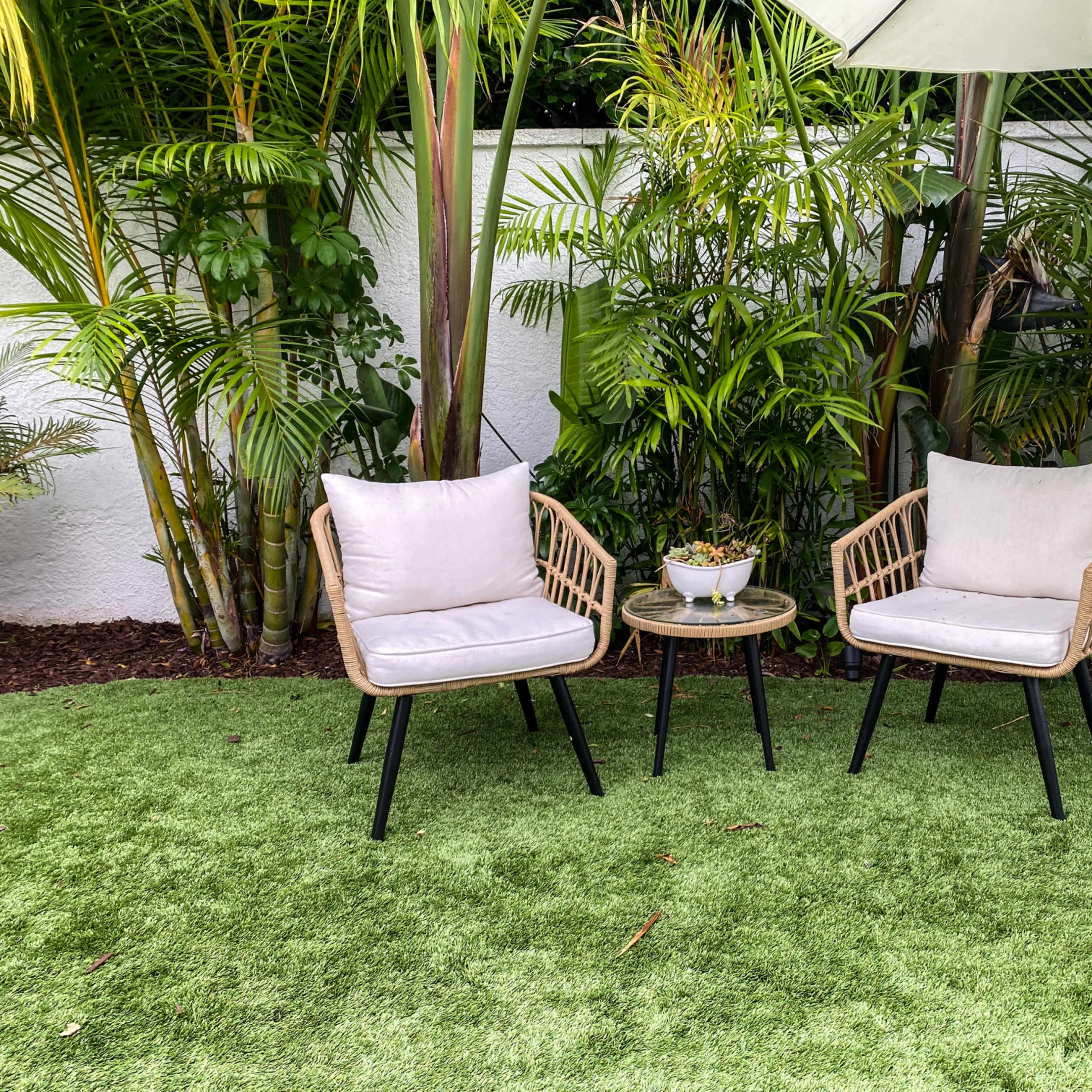 Two wicker chairs with light cushions sit beside a small table on artificial grass, surrounded by lush greenery.