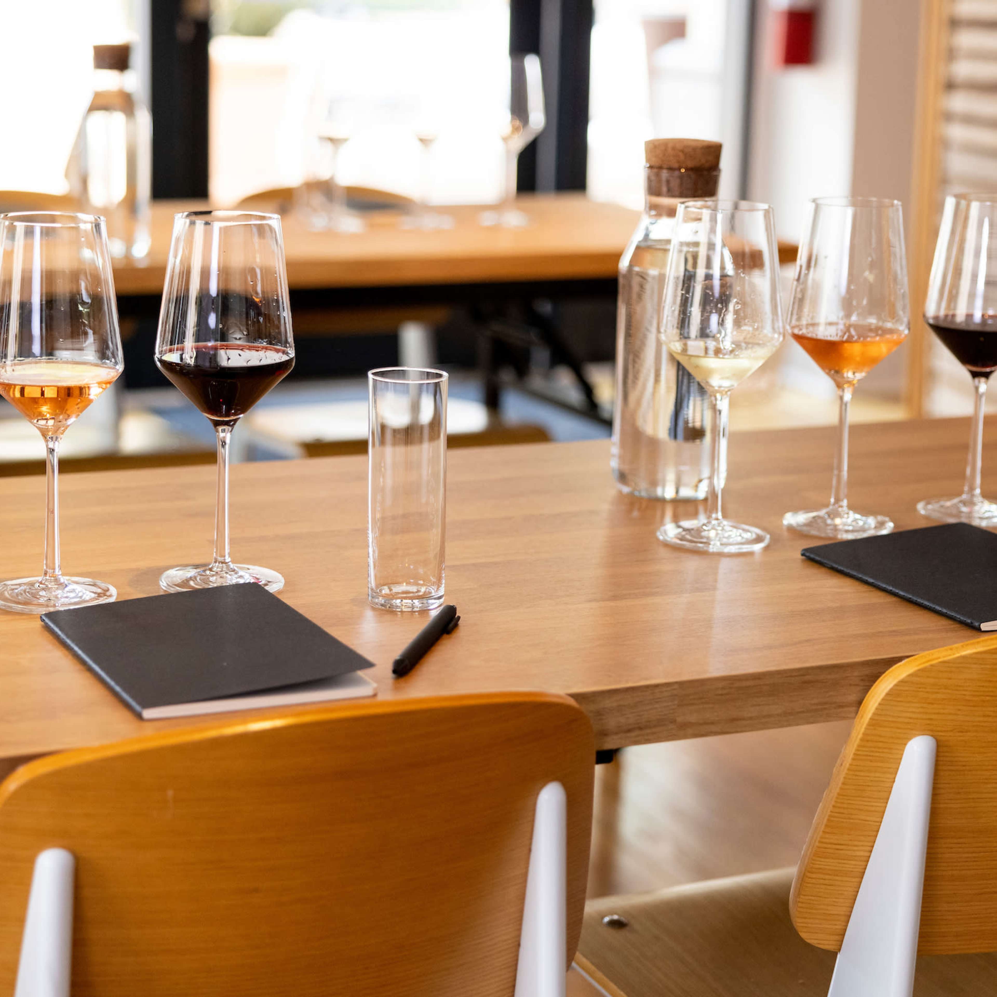 A wooden table is set with several wine glasses and a water bottle, along with notepads and pens for each place setting.