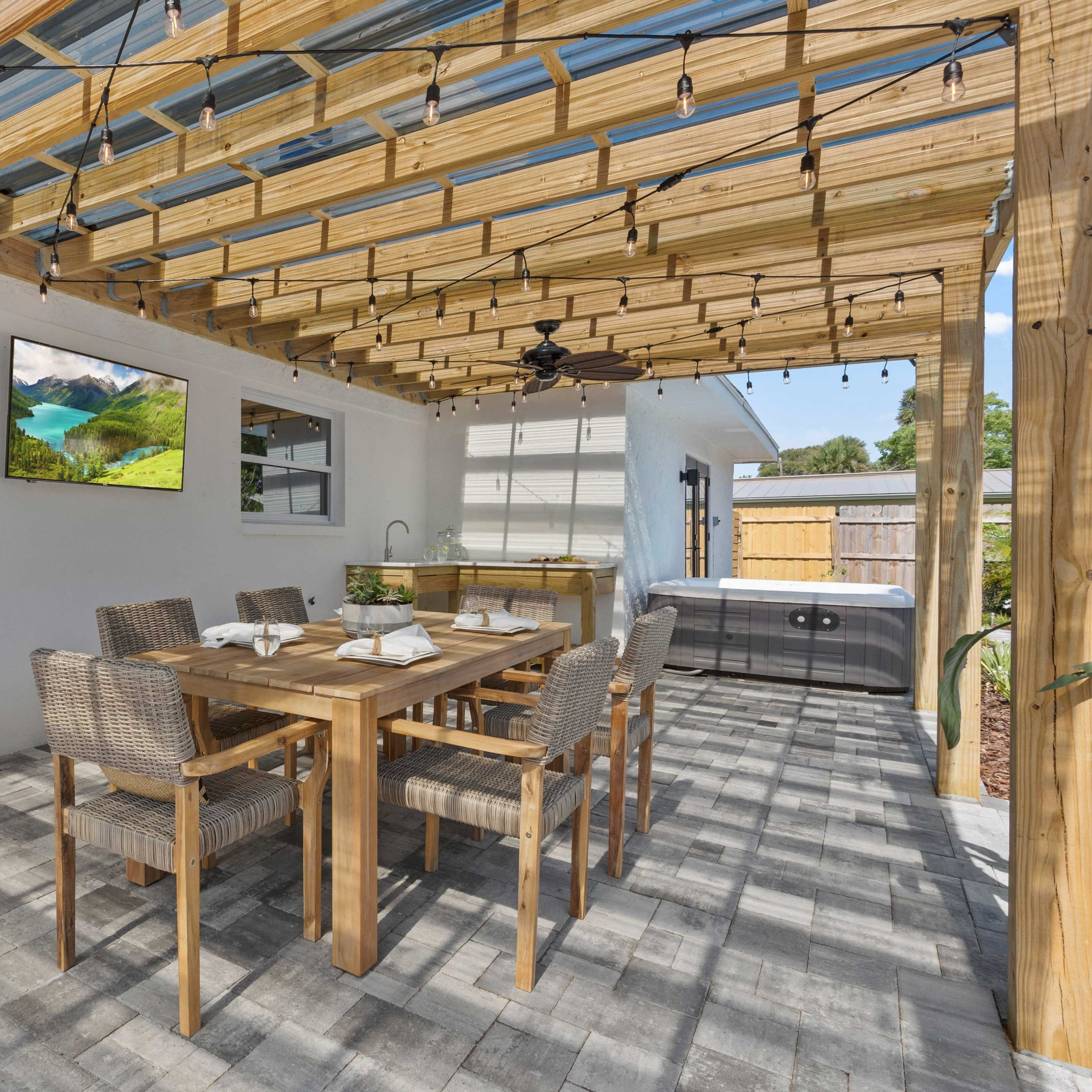 A covered outdoor dining area with a wooden table and chairs, a mounted TV, and a hot tub in a well-lit patio setting.