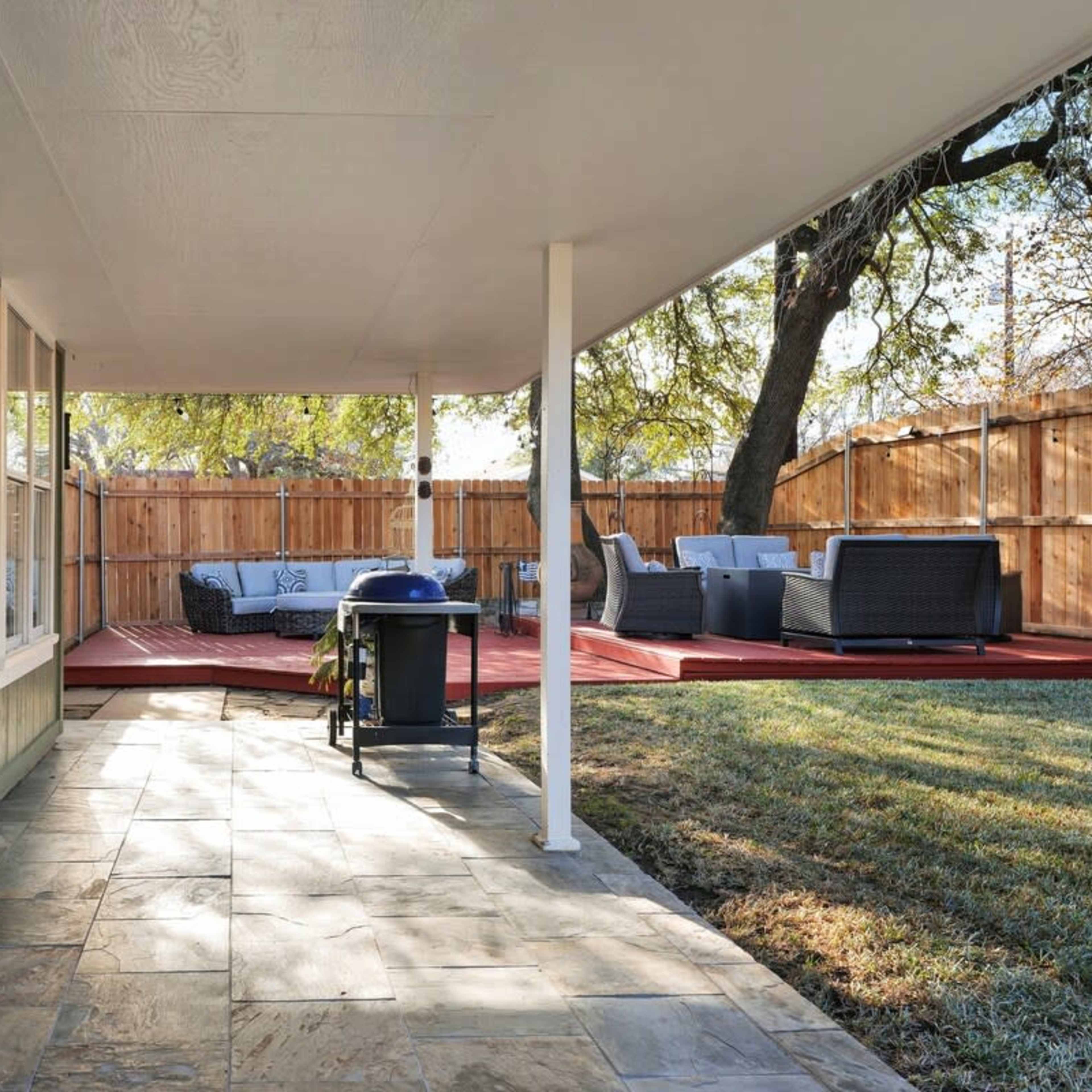 A covered patio with a stone floor leads to a wooden deck and a backyard, featuring a grill and outdoor seating.