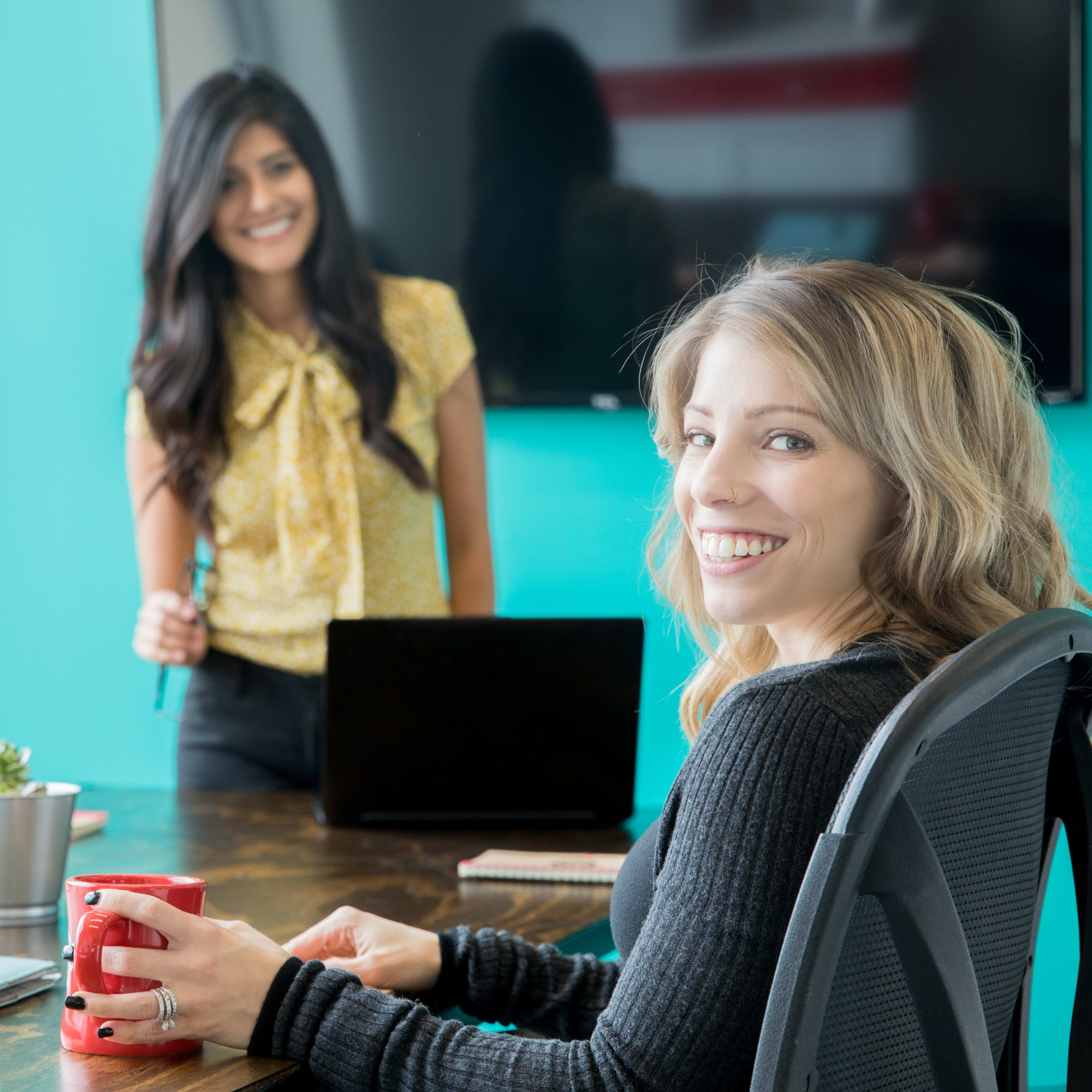 Two women are seated at a conference table, with one standing in the background and the other smiling at the camera while holding a red mug.