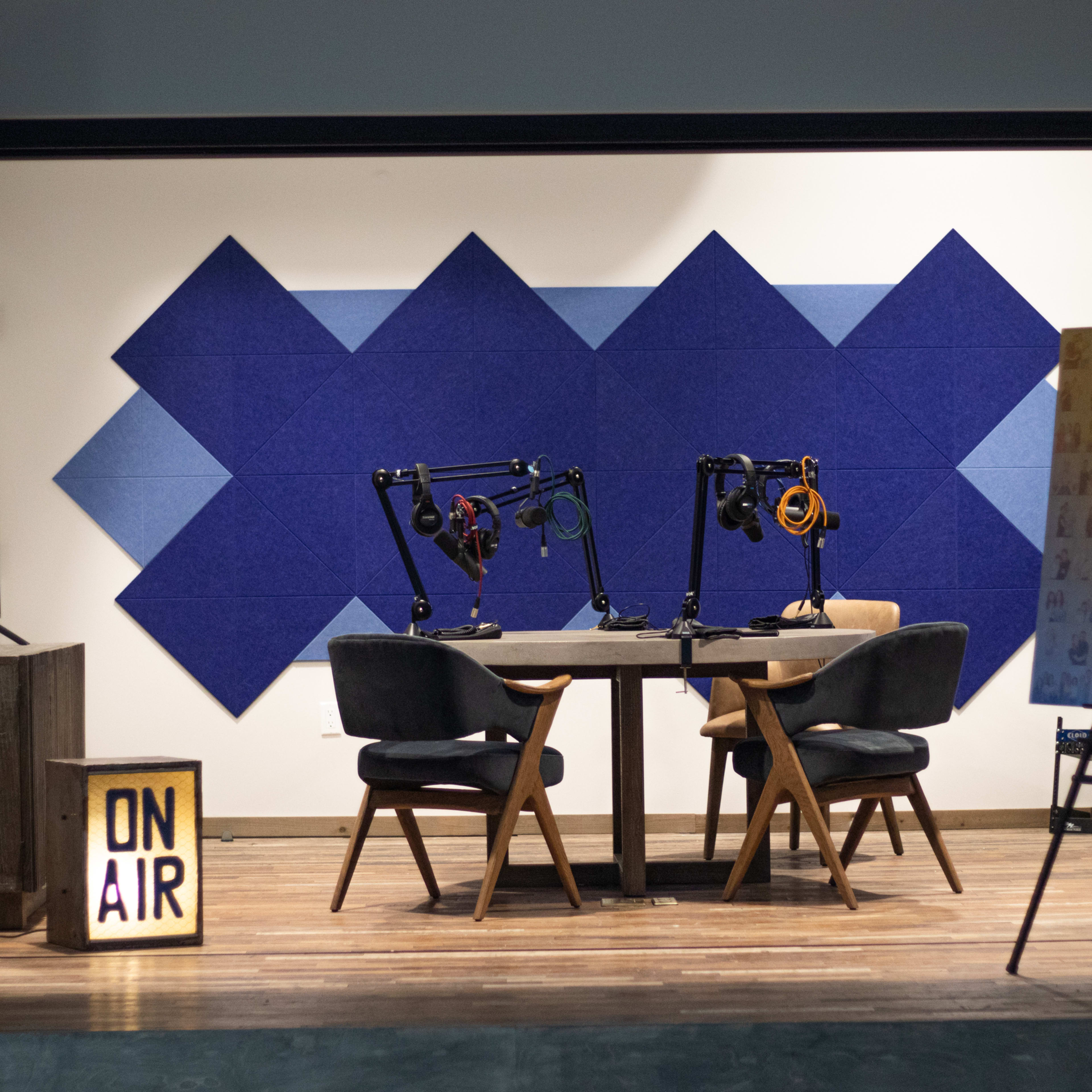 A podcast recording studio featuring a wooden table with two chairs, microphone arms, soundproof blue wall panels, and an "ON AIR" sign.