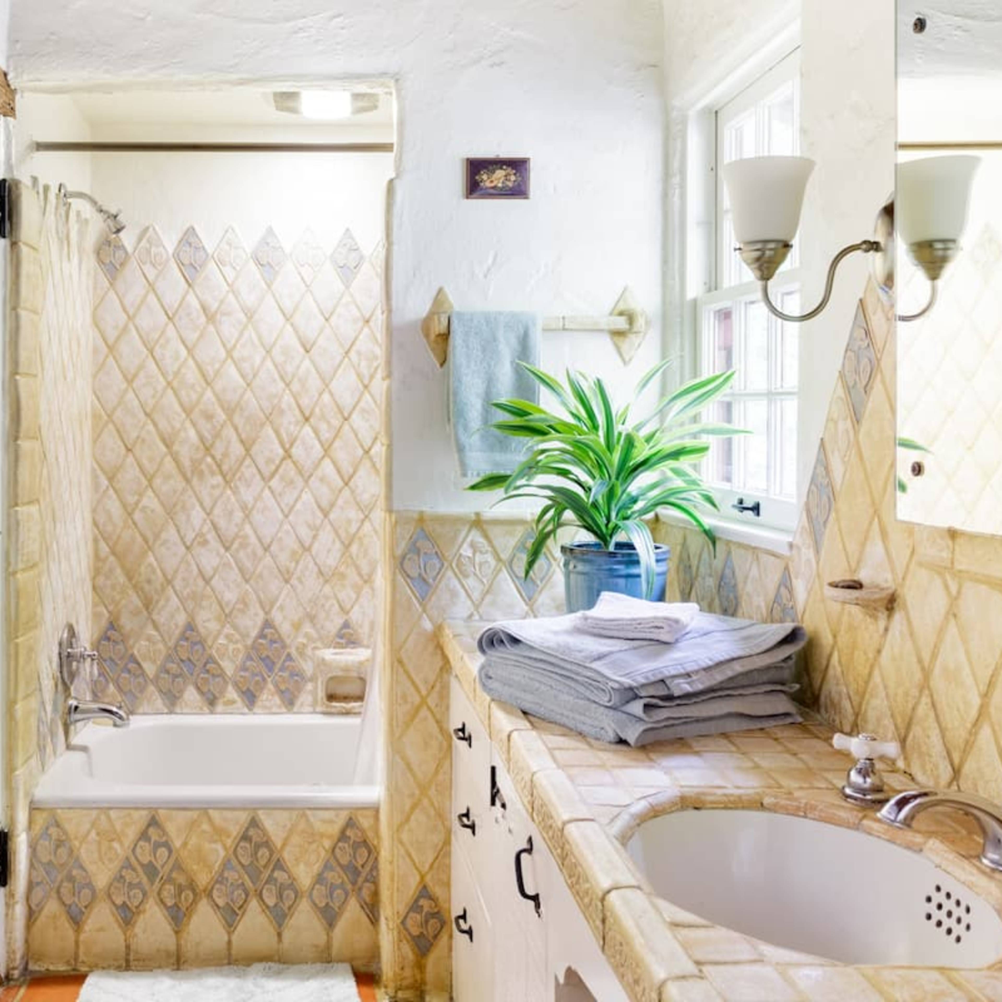 A well-lit bathroom features a bathtub with tiled walls, a sink with a large mirror, and a potted plant on the countertop.