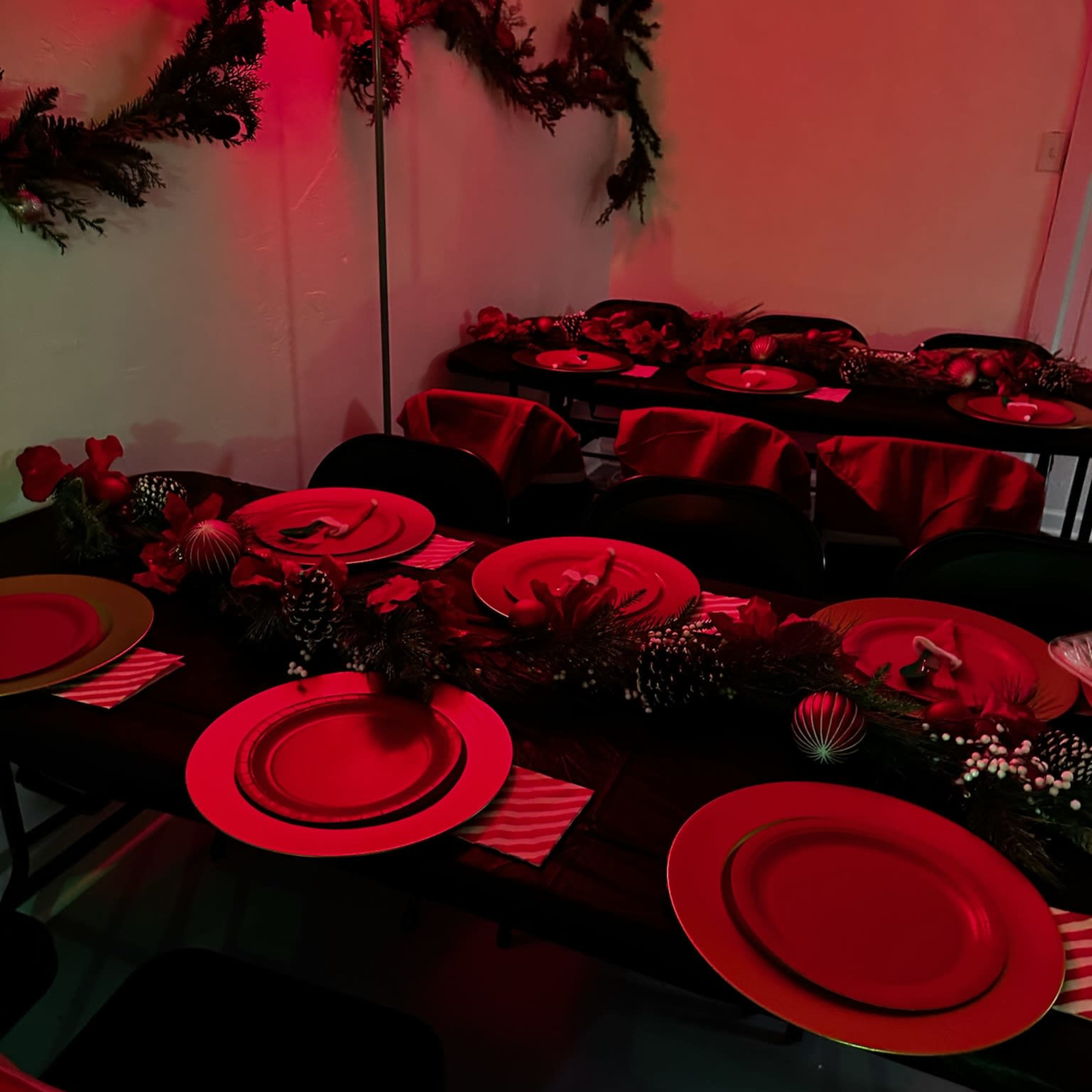 A dining area is set with red plates and decorations, featuring a long table adorned with garlands and floral accents under red lighting.