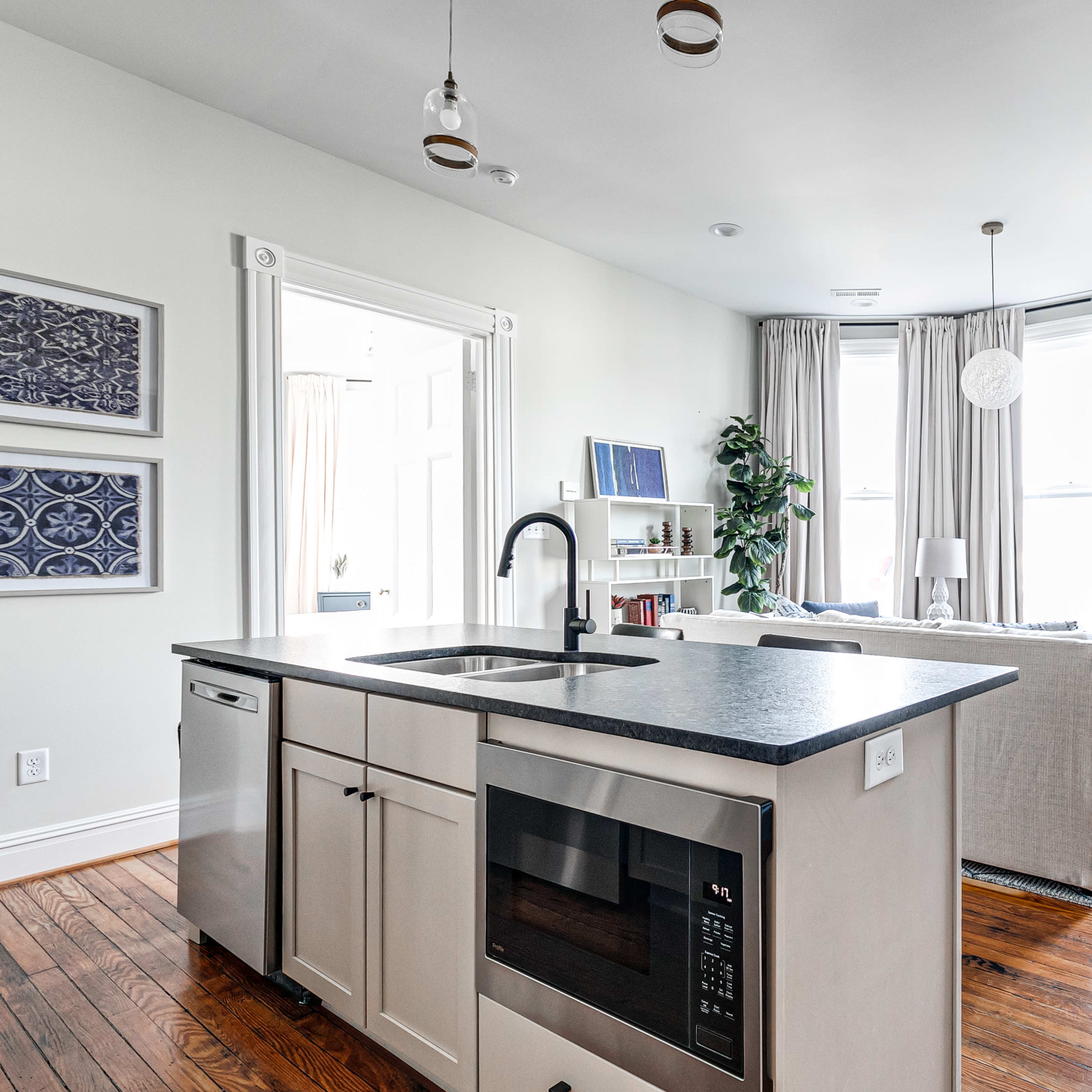 A modern kitchen features a central island with a microwave, overlooking a bright living area with large windows and light-colored curtains.