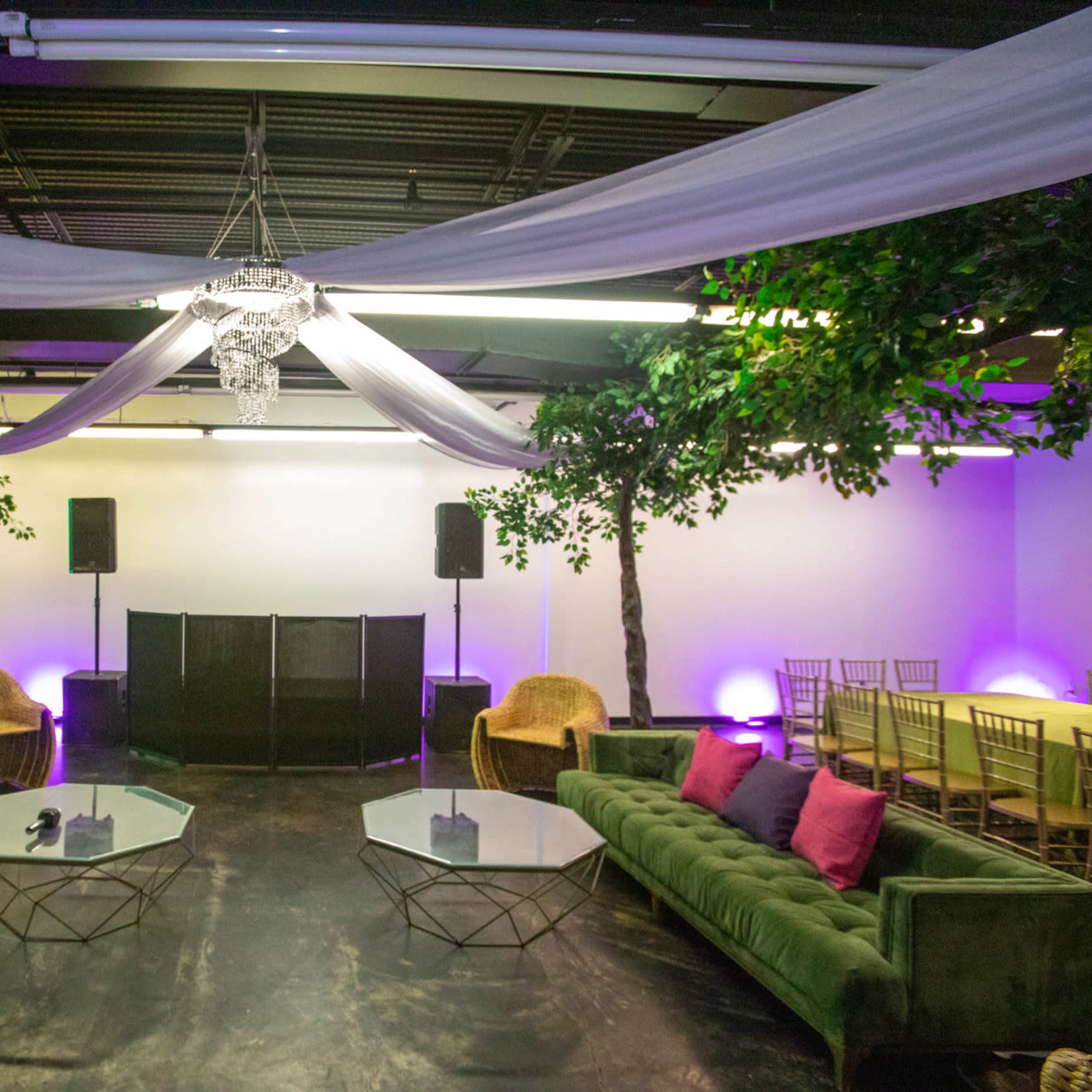 A spacious event venue with green sofas, glass coffee tables, and a stage backdrop adorned with trees and draped fabric.