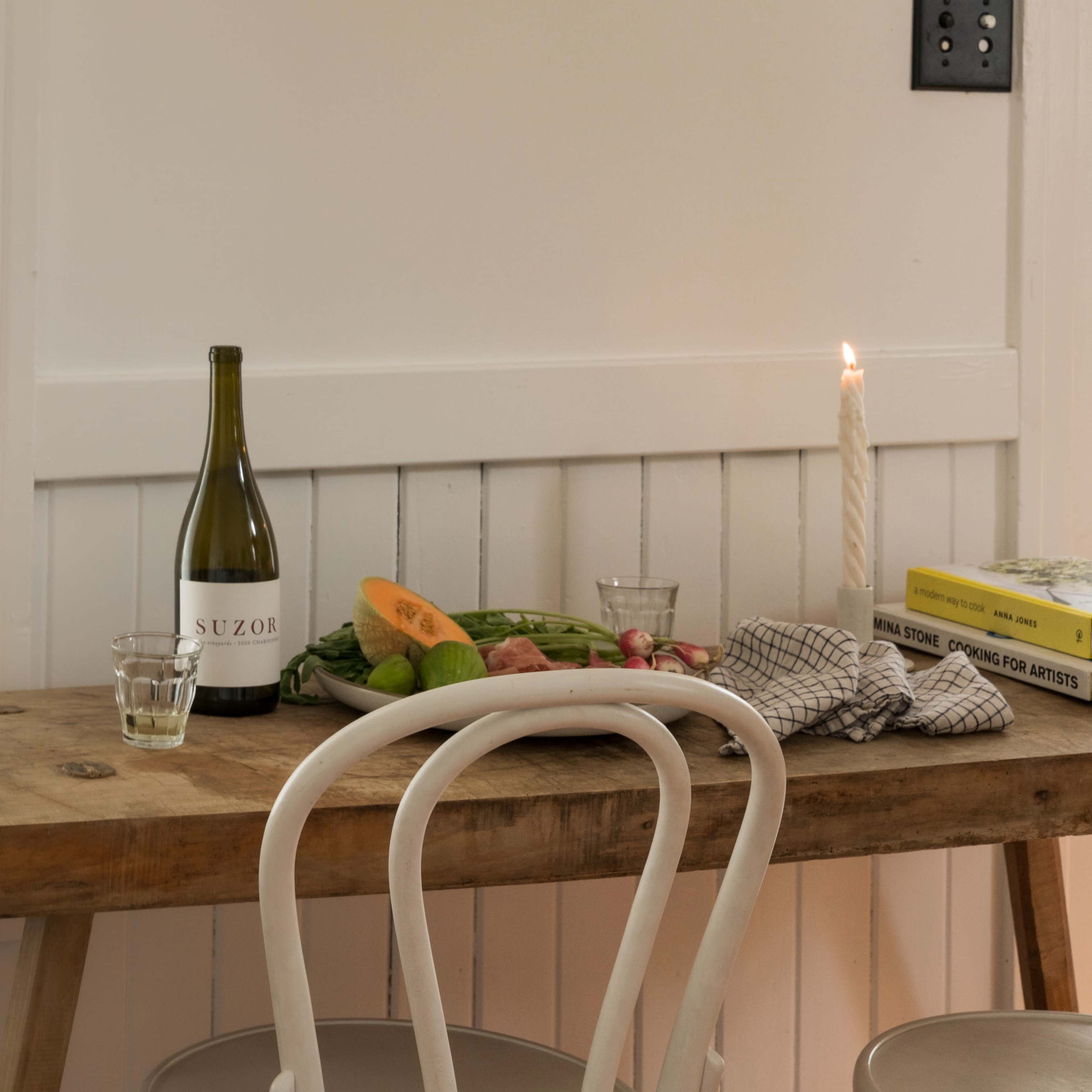 A wooden table is set with a bottle of wine, fresh fruits and vegetables, a candle, and a stack of books.