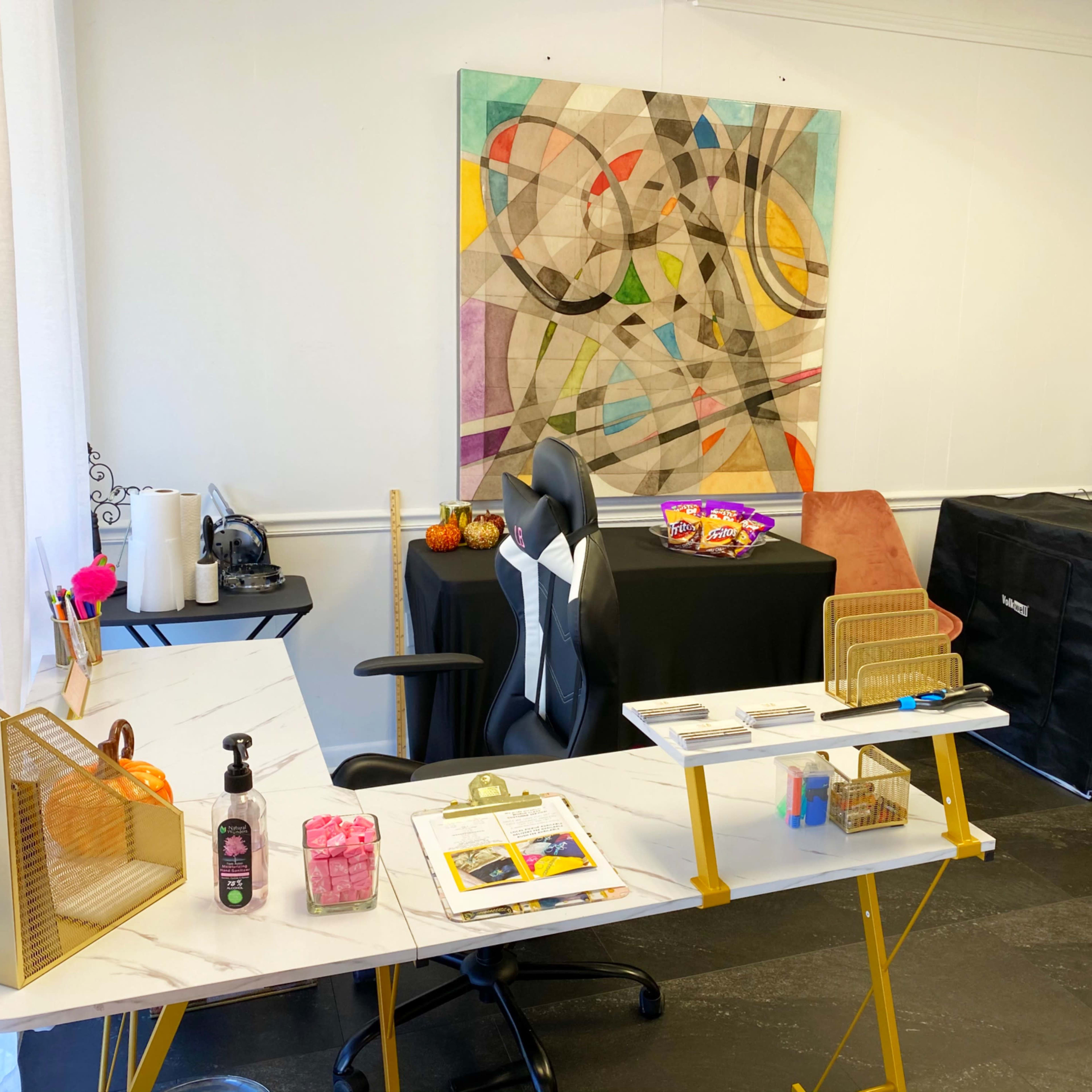 A well-organized room featuring a modern desk with a chair, a large abstract painting on the wall, and various items displayed on a black table and a grid panel.
