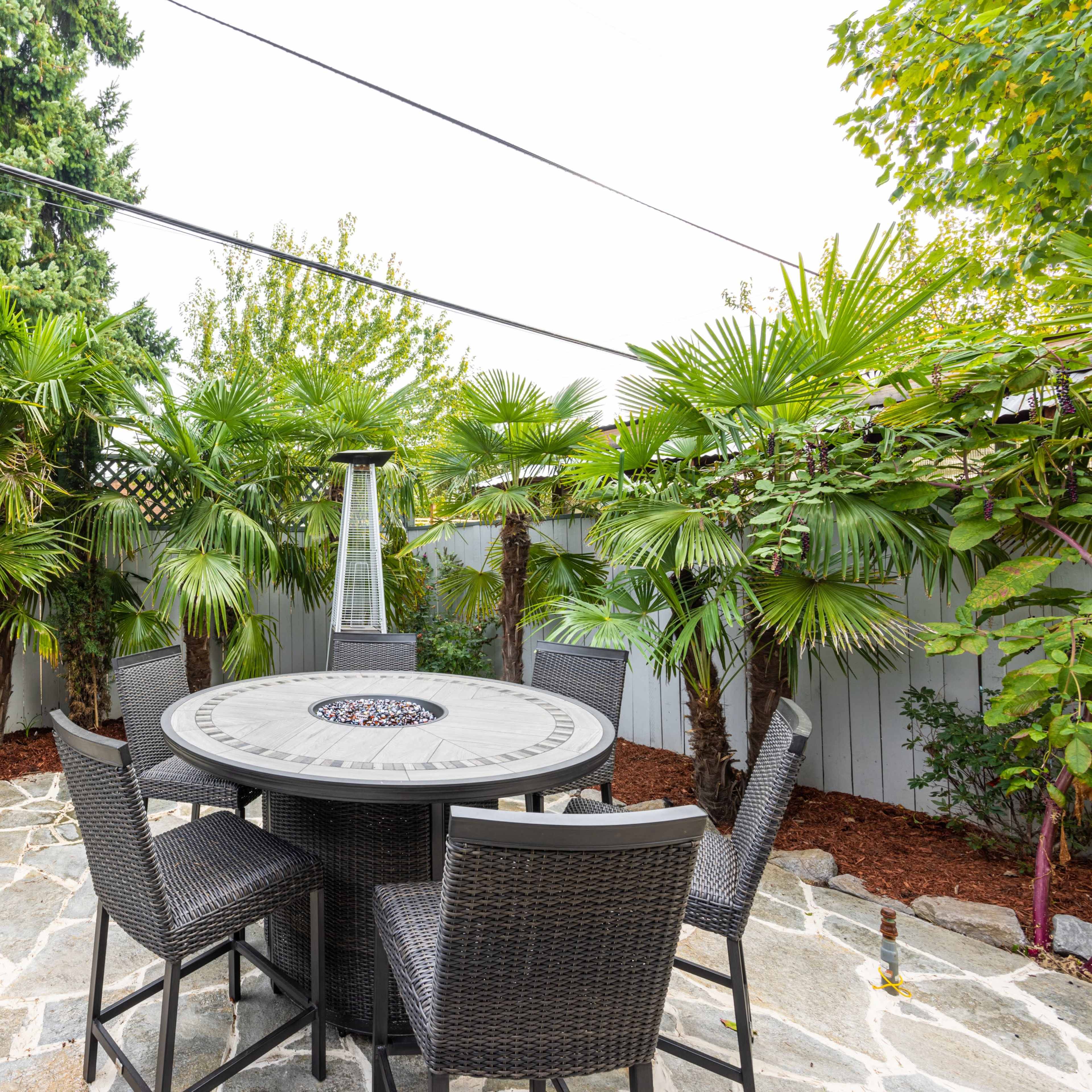 A circular stone patio table surrounded by eight chairs is set among tall palm trees and outdoor heaters in a landscaped yard.