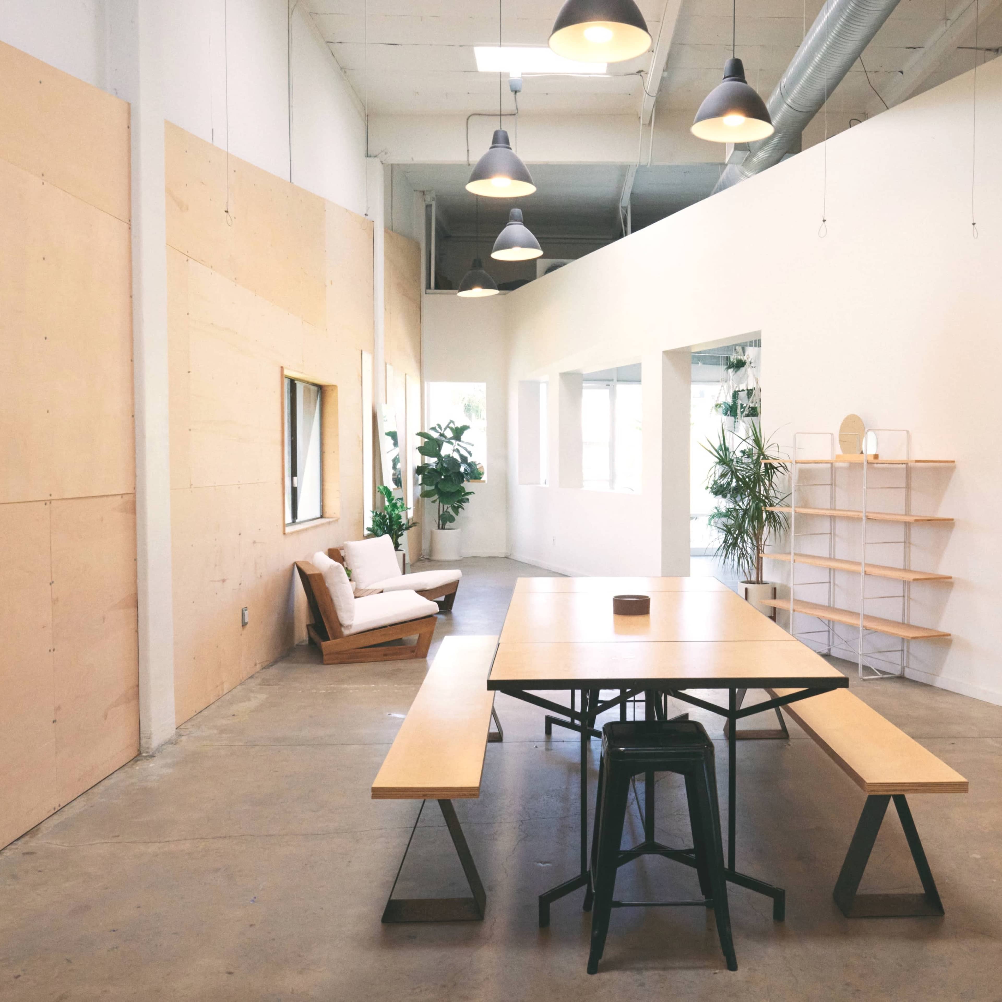 A spacious, well-lit room features a wooden table with benches, surrounded by indoor plants and minimalist decor.