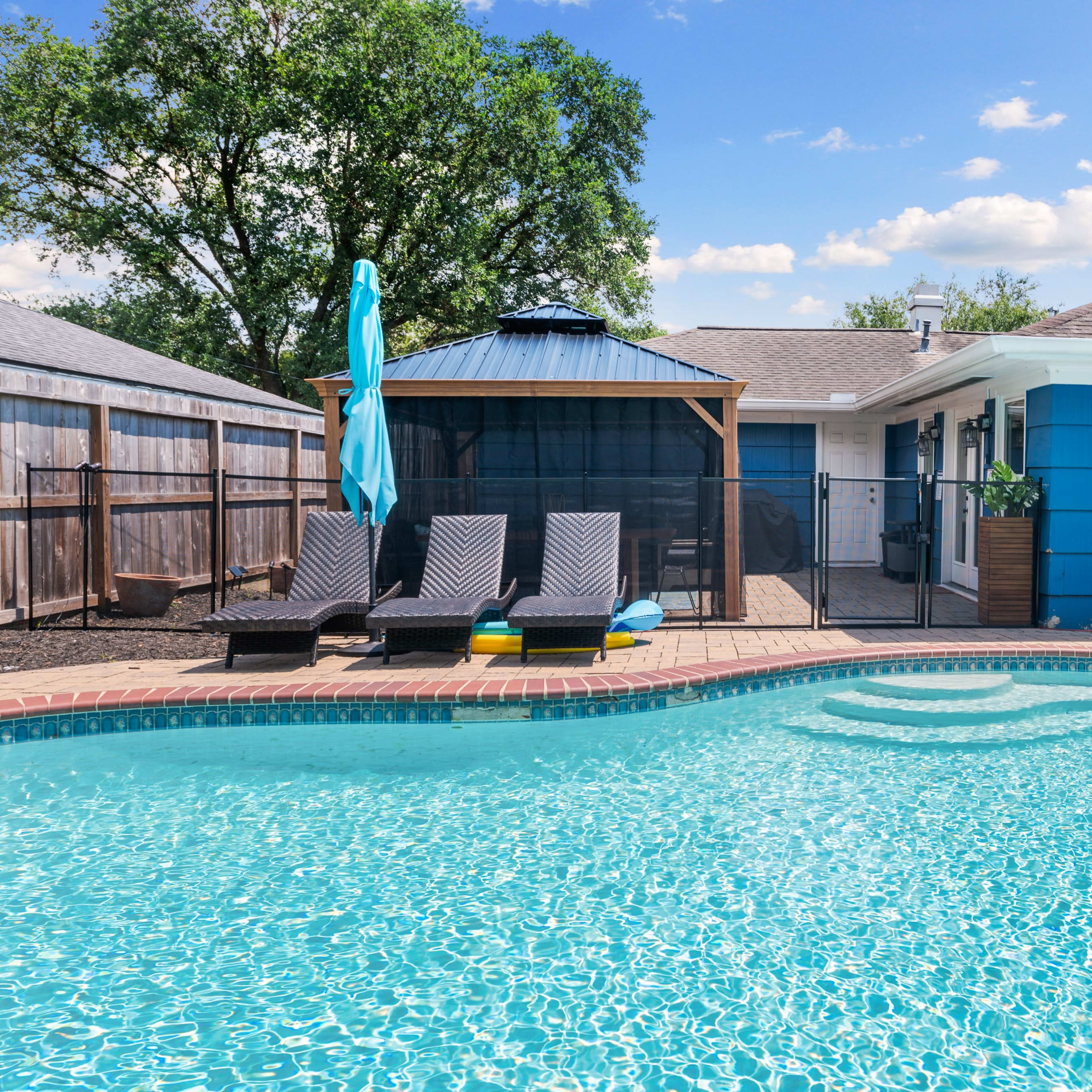 A backyard with a swimming pool, two lounge chairs, and a gazebo beside a blue house.