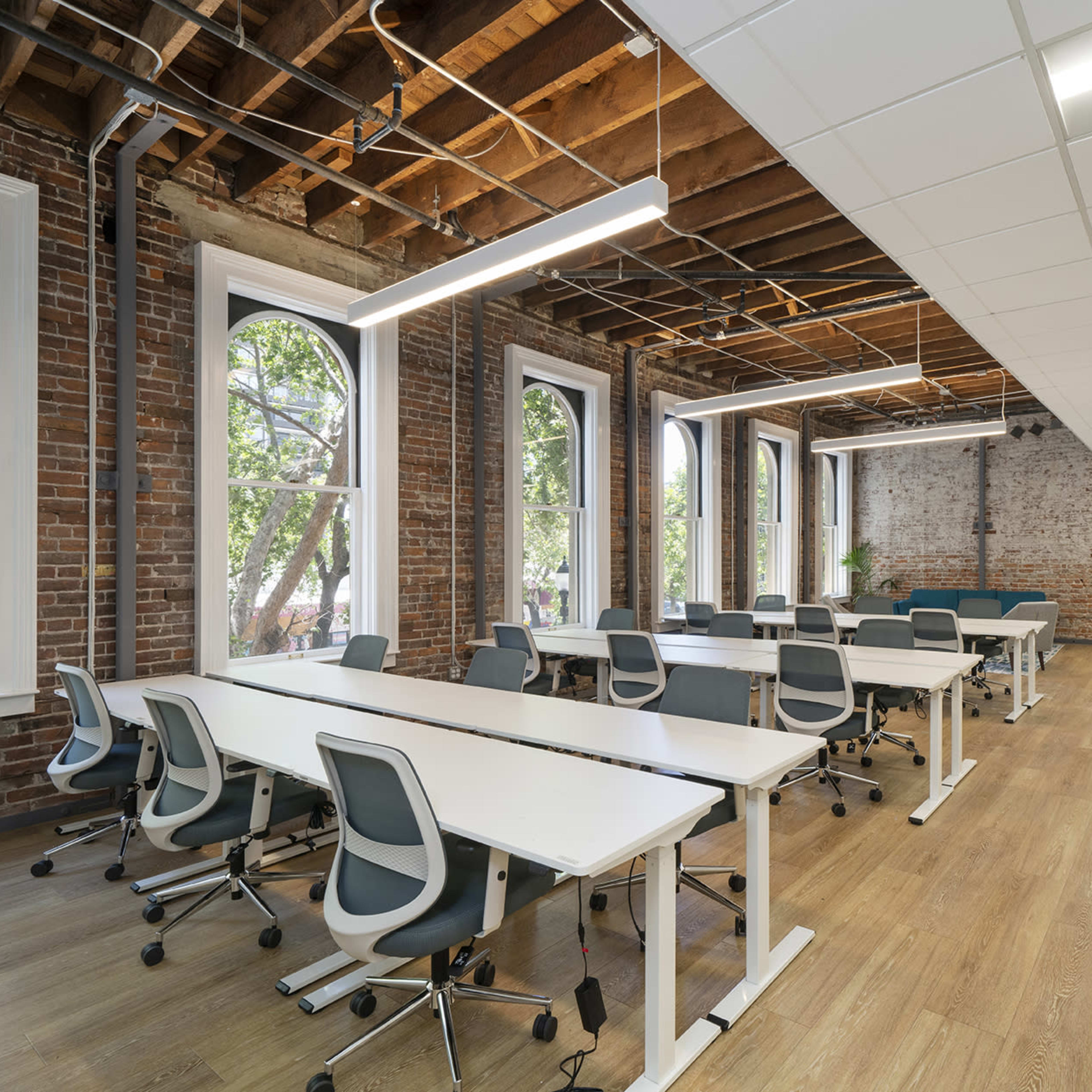 A modern office space features multiple workstations arranged in a bright room with large windows and exposed brick walls.