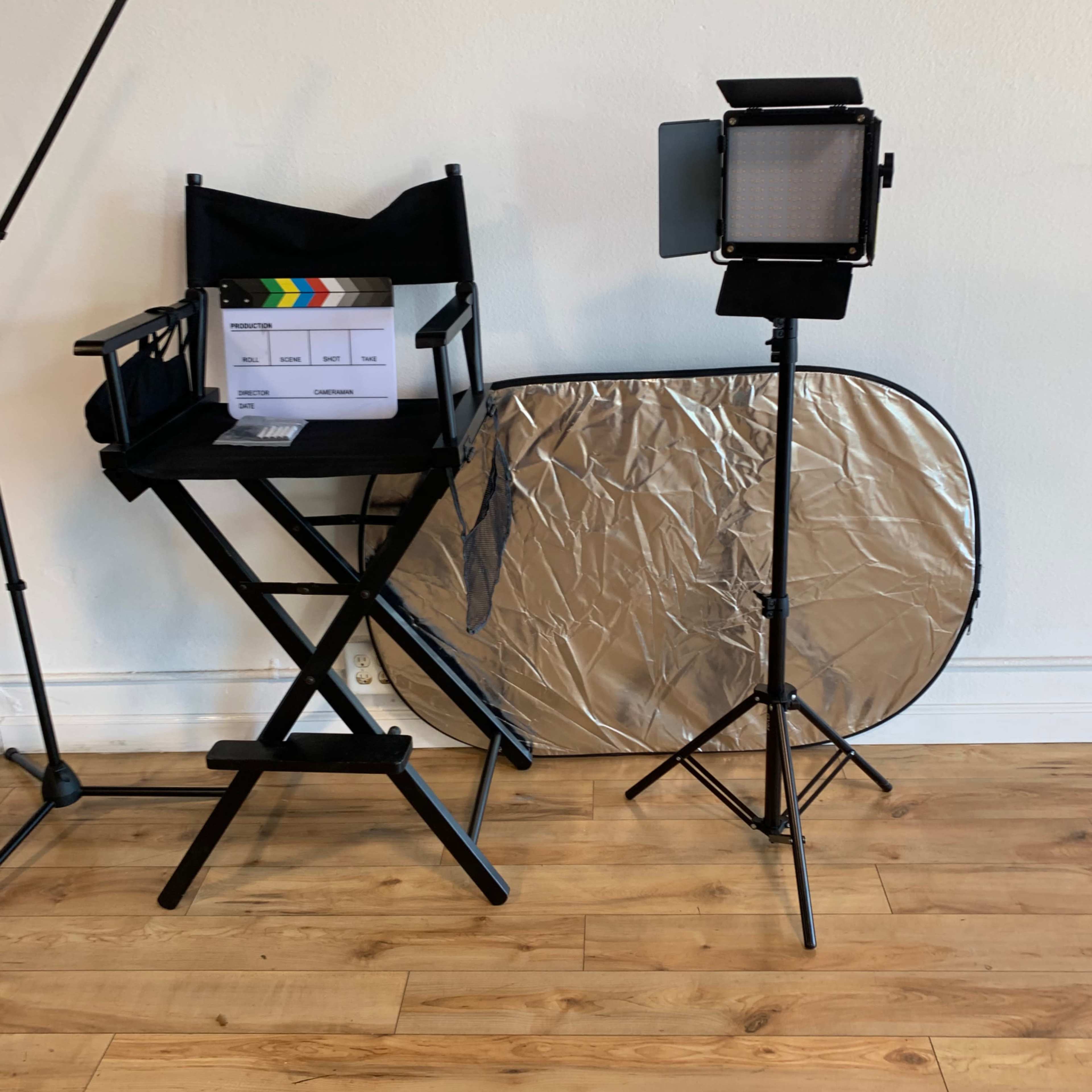 A director's chair, a clapperboard, a lighting setup, and a reflective disc are arranged on a wooden floor against a plain wall.