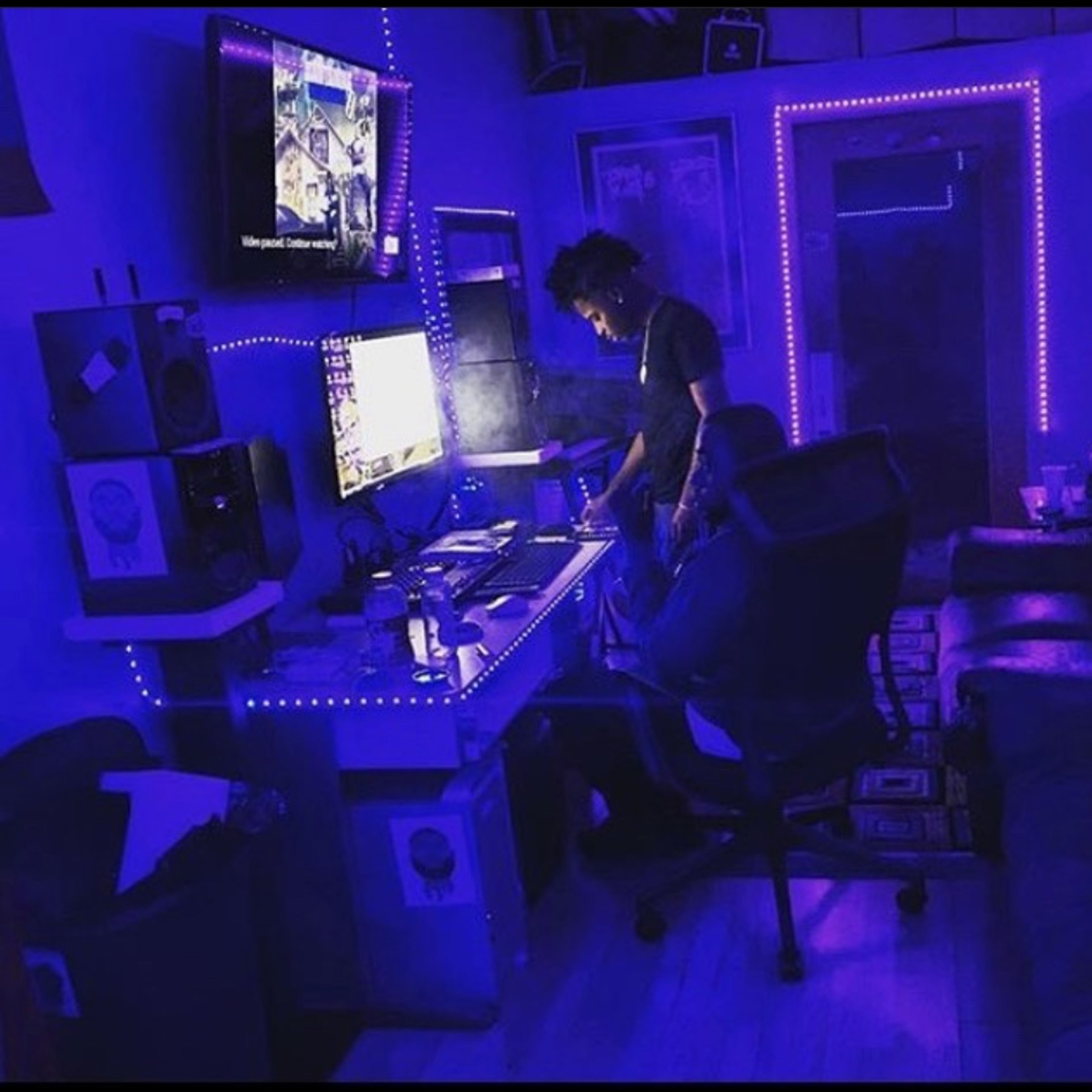 A person stands at a desk with multiple monitors in a room illuminated by blue LED lights, while another seated figure is partially visible on a couch.