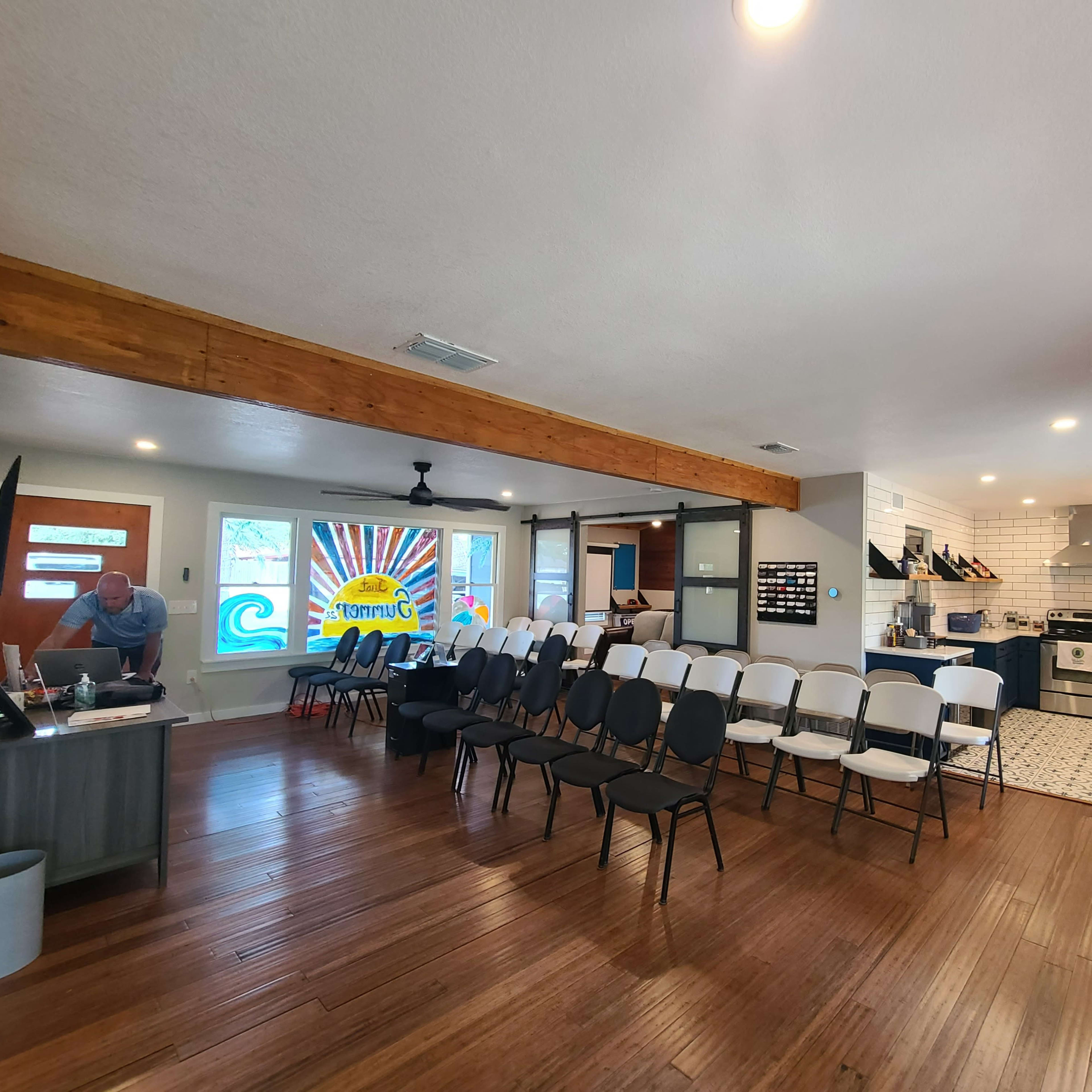 A spacious room with wooden flooring, featuring rows of chairs, a modern kitchen area, and colorful wall decor.