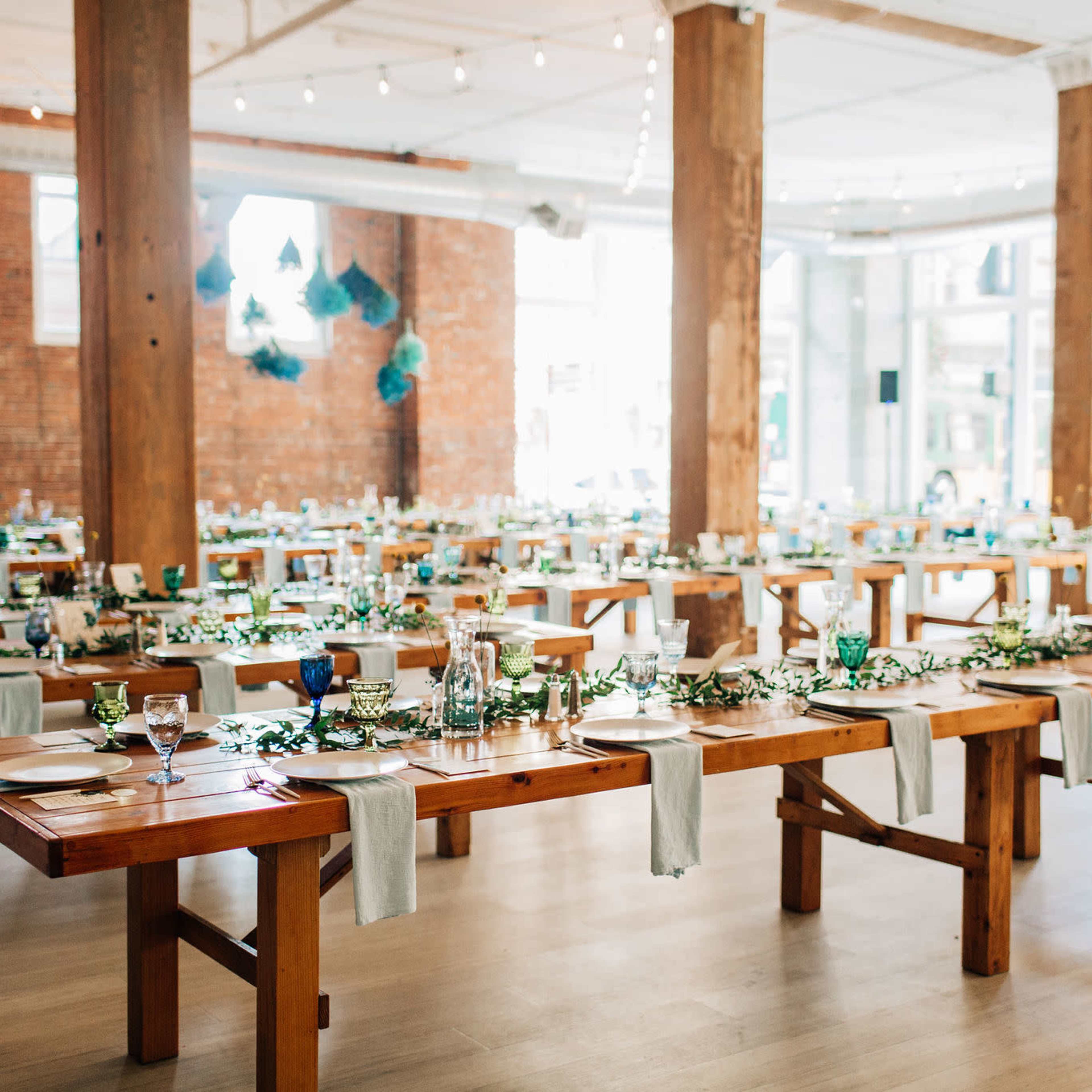 A spacious event venue with numerous wooden tables arranged for dining, featuring blue and green glassware and floral centerpieces.