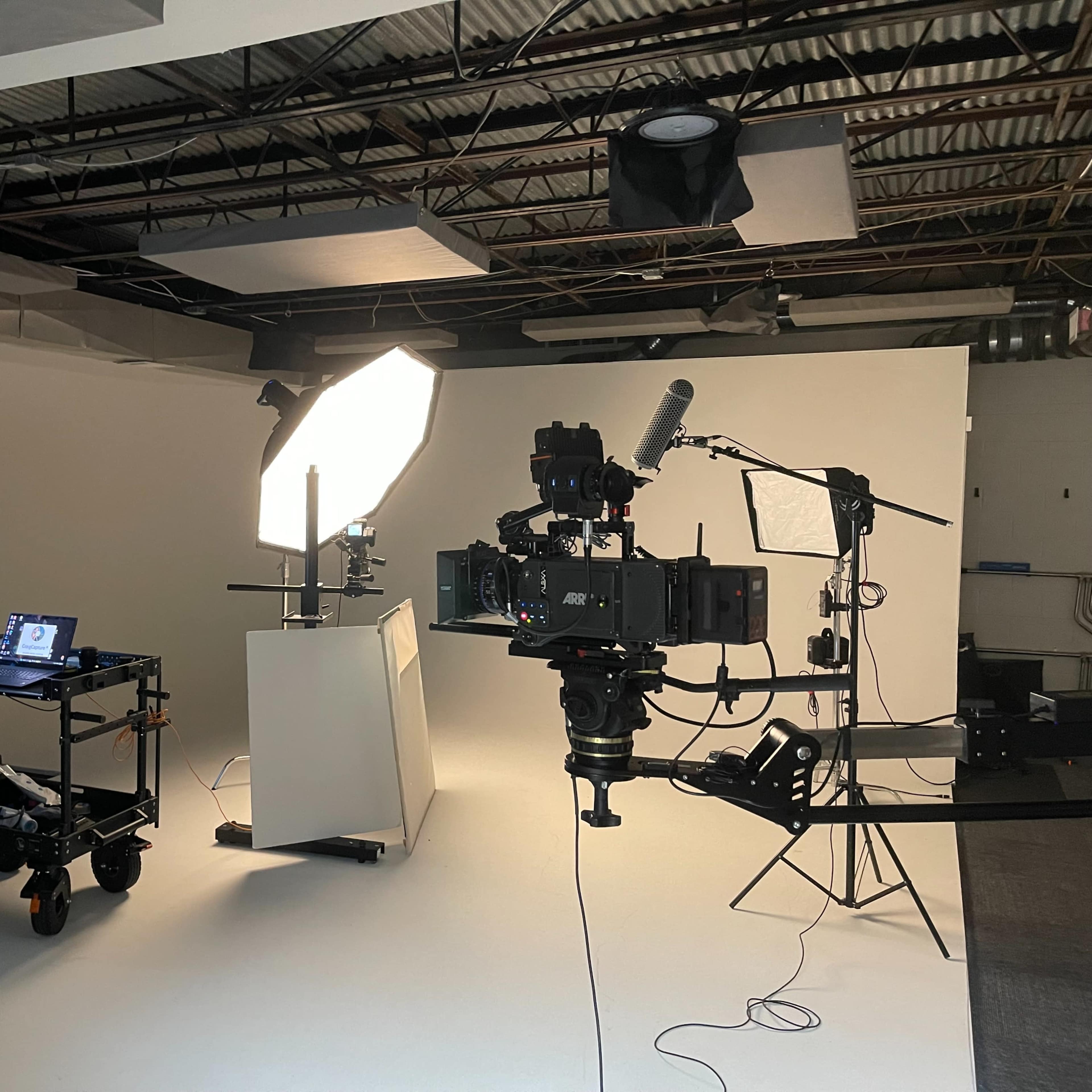 A video production setup features a camera on a tripod, an illuminated backdrop, a computer display on a cart, and various lighting equipment in a studio environment.