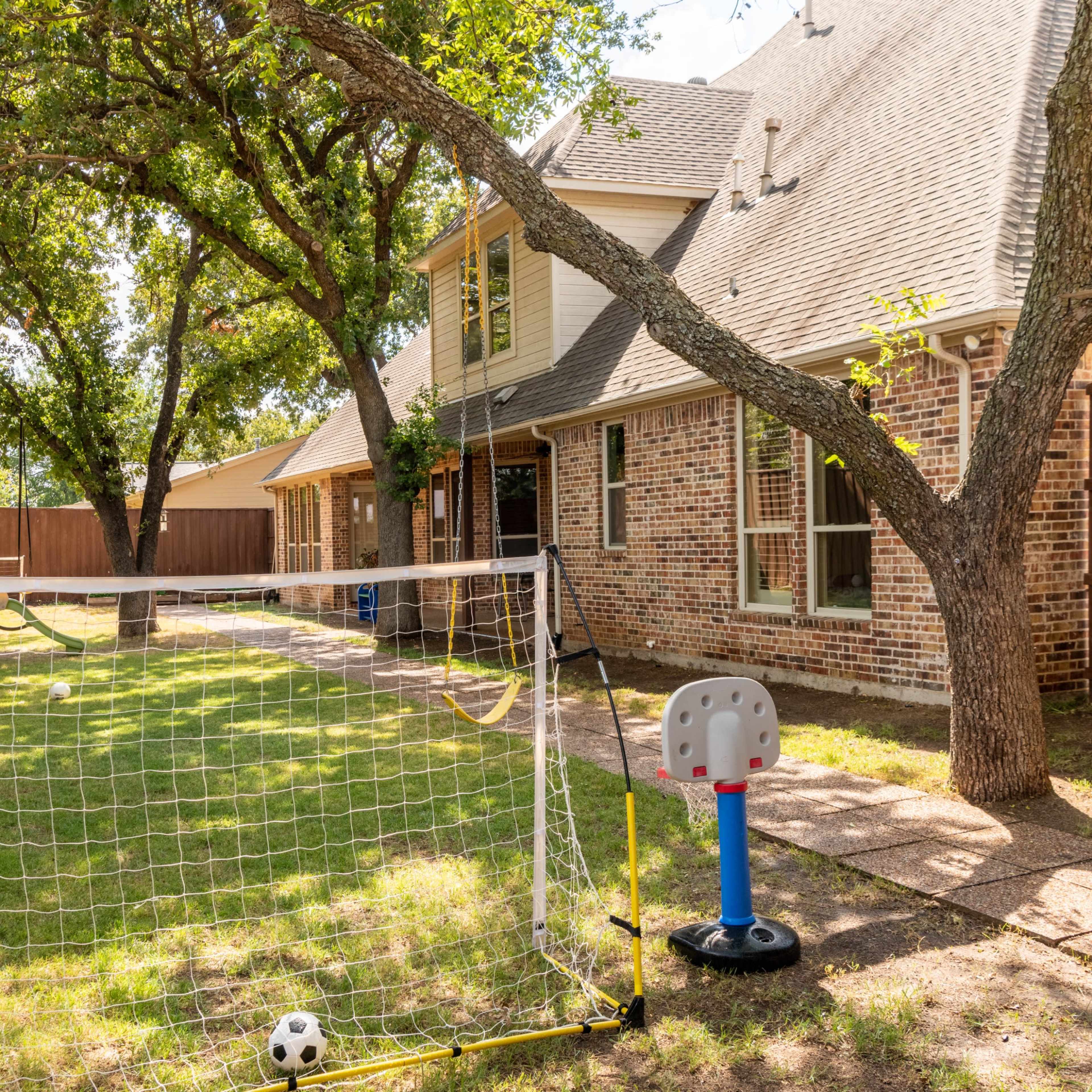A backyard with a soccer goal, a small basketball hoop, and a brick house in the background surrounded by trees and a wooden fence.